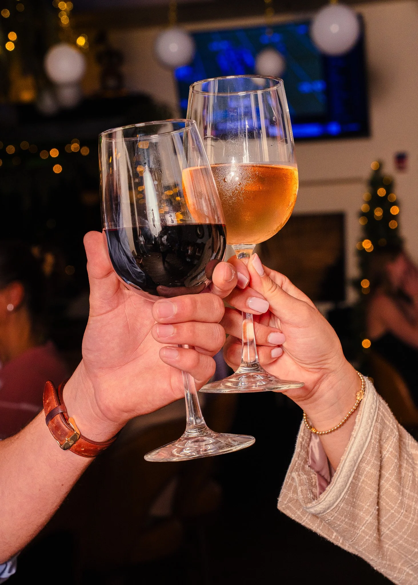 Two people clinking glasses, one with red wine and the other with rose, in a festive setting.