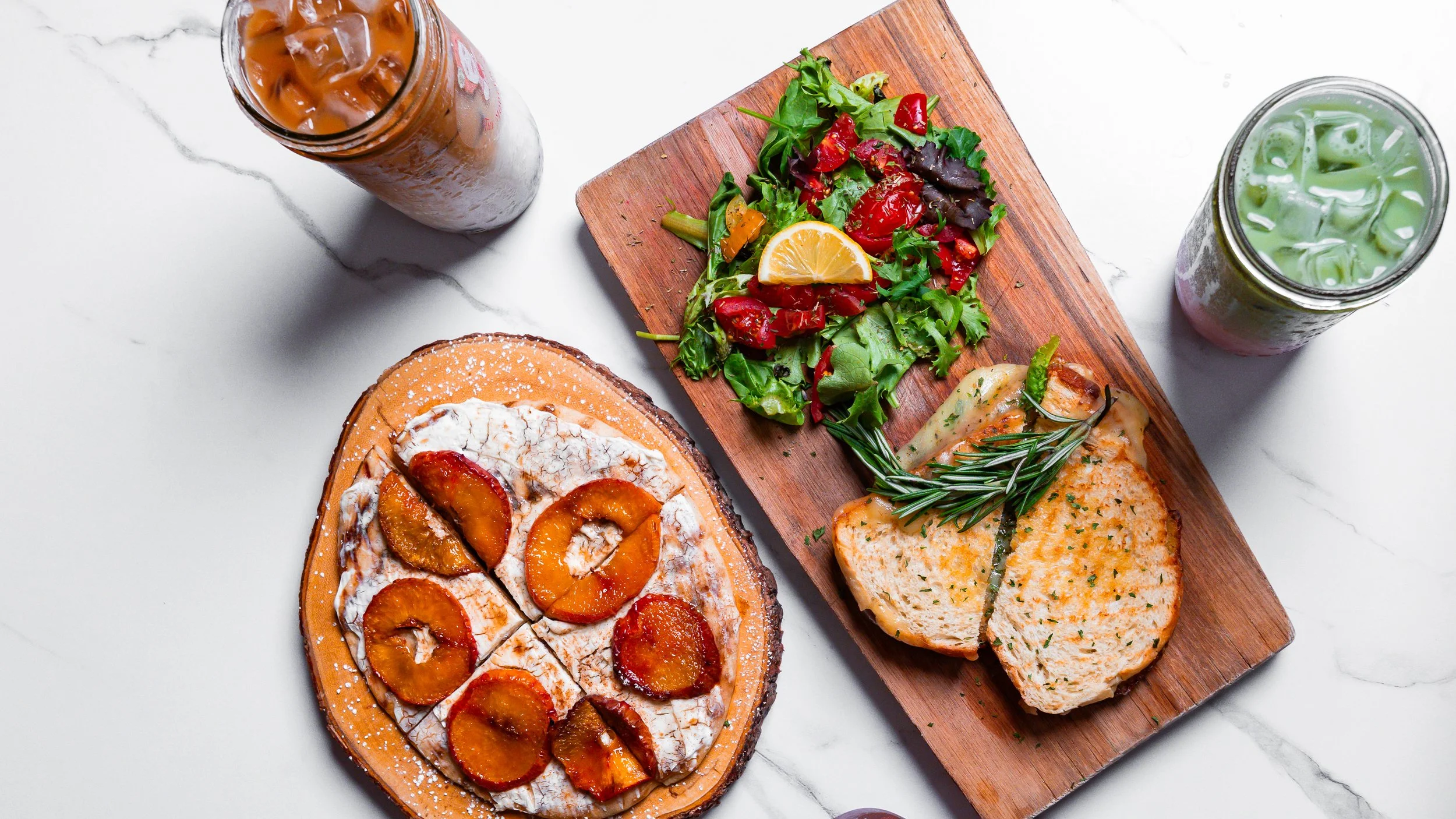 A marble table with two beverages, a cutting board with salad and toasted sandwiches, and a dessert pizza topped with caramelized fruit.