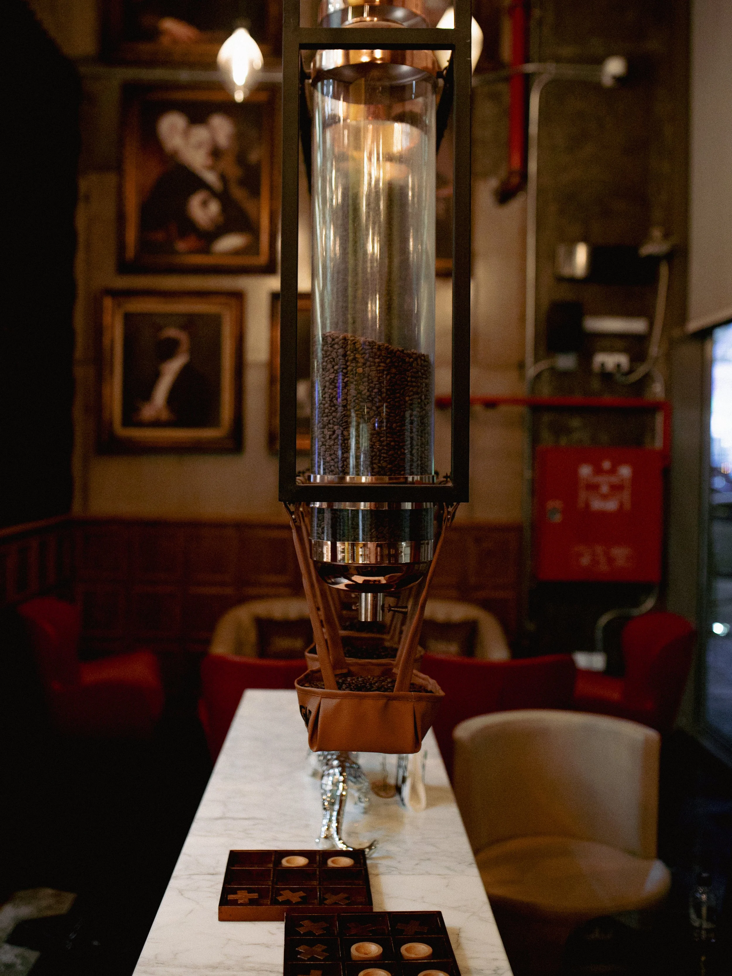Large glass coffee bean dispenser in a cafe with decorative paintings and lighting in the background.