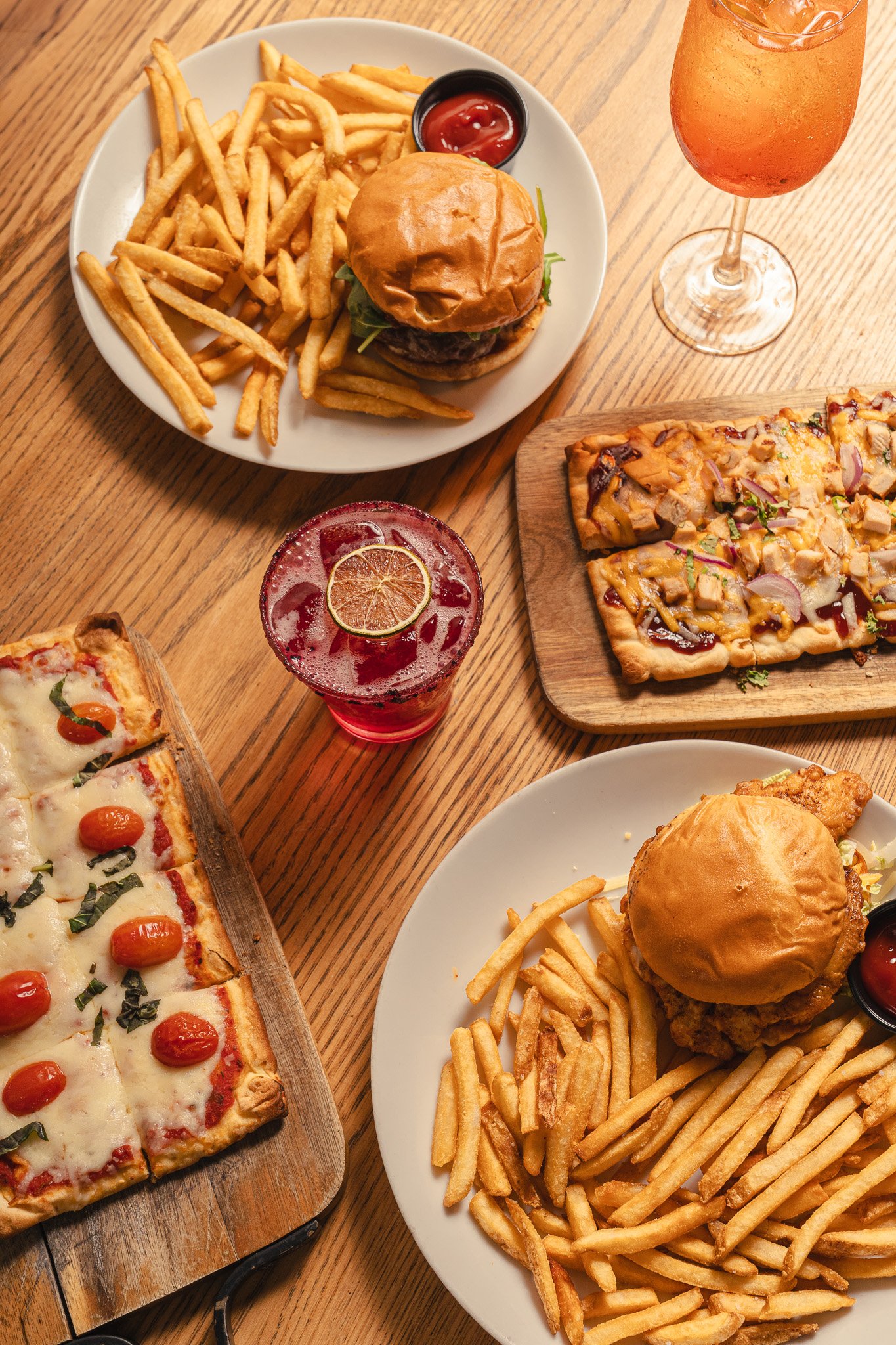 Overhead view of pizza, burgers with fries, and cocktails on a wooden table.