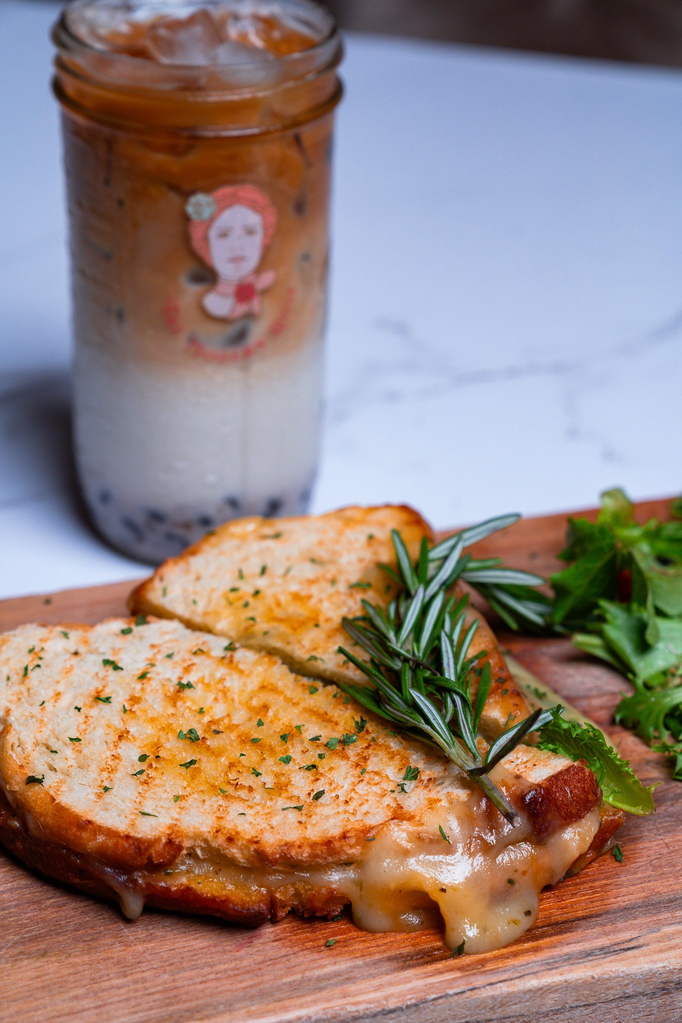 Grilled cheese sandwich on wooden board with rosemary garnish, side salad, and iced latte in a jar on a table.