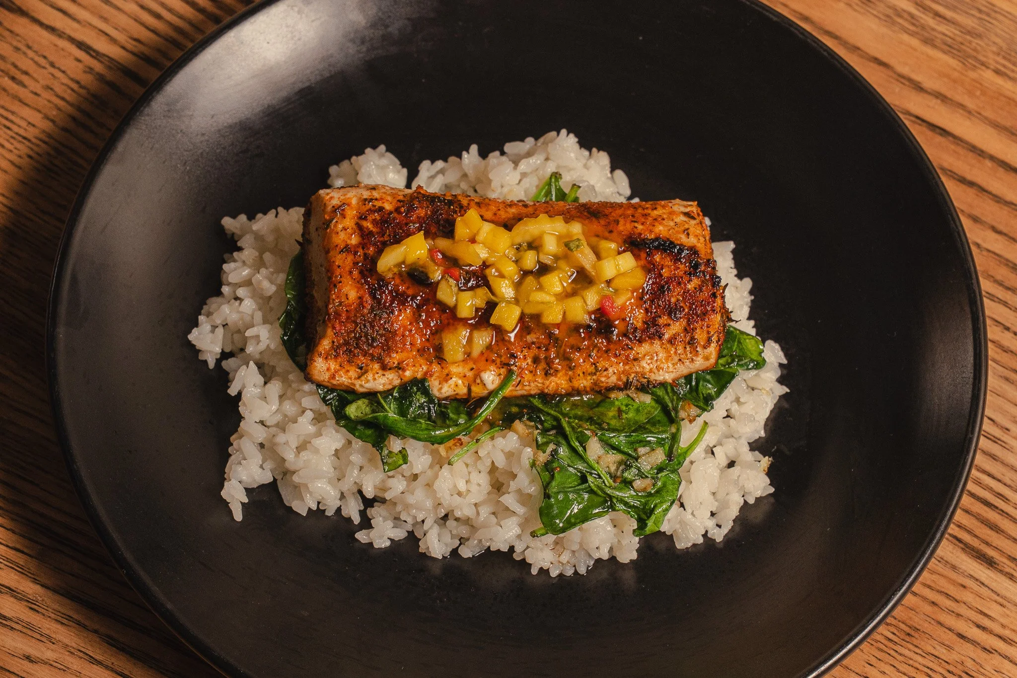 Grilled salmon with mango salsa on spinach and rice, served on a black plate.
