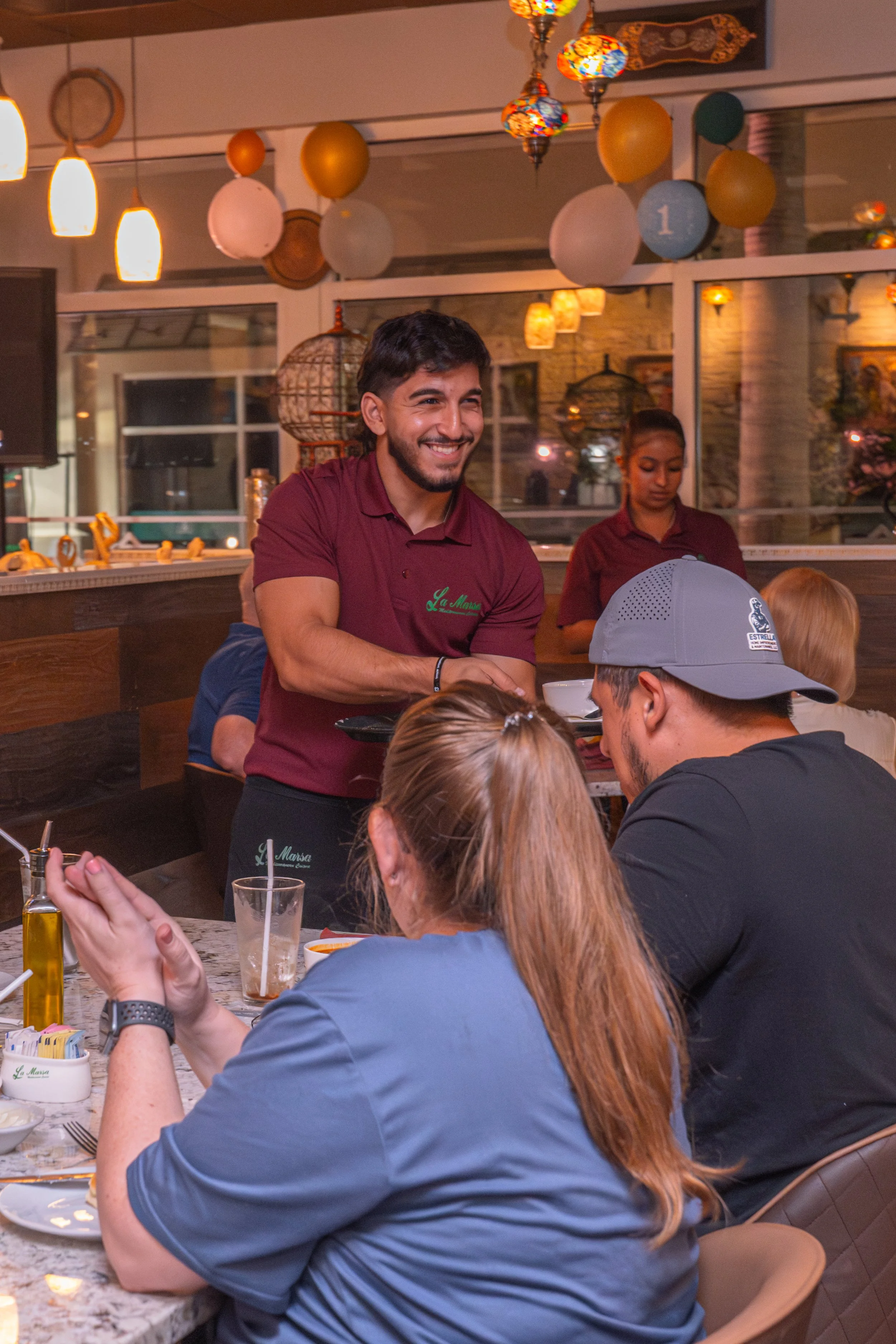 Restaurant scene with a smiling waiter serving a table of patrons, with balloons and decorative lights in the background.