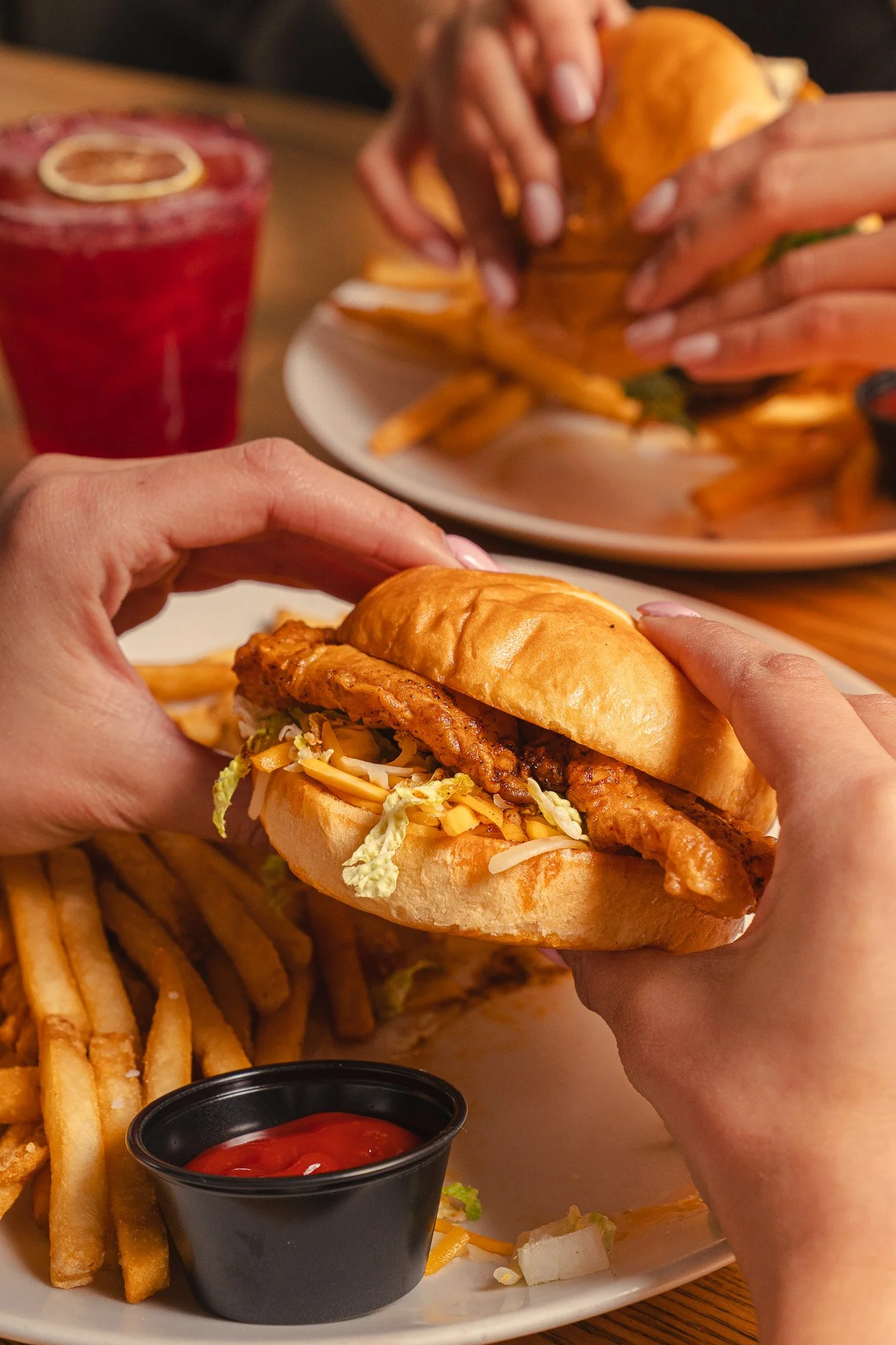 Close-up of hands holding a chicken sandwich with fries and ketchup on a plate, and a red drink in the background.