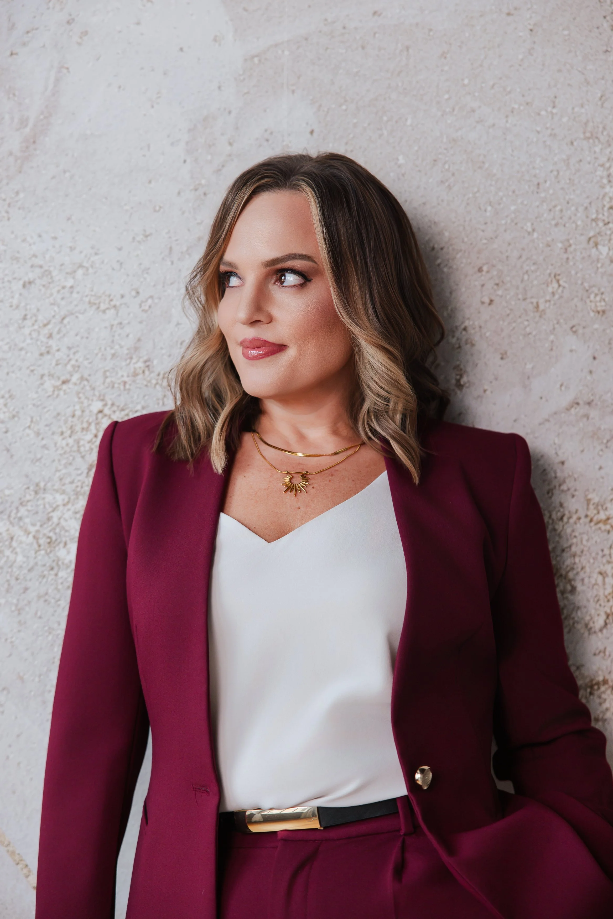 Woman with shoulder-length wavy hair, wearing a burgundy blazer over a white blouse, standing against a textured light-colored wall.