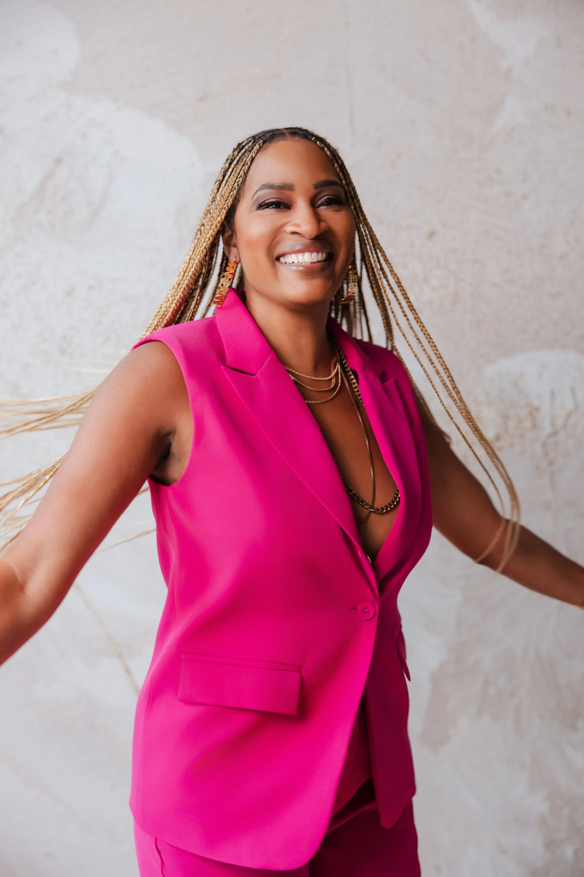 Woman smiling, wearing a bright pink sleeveless blazer and matching pants, with blonde braided hair, gold jewelry, and earrings, standing against a light textured background.