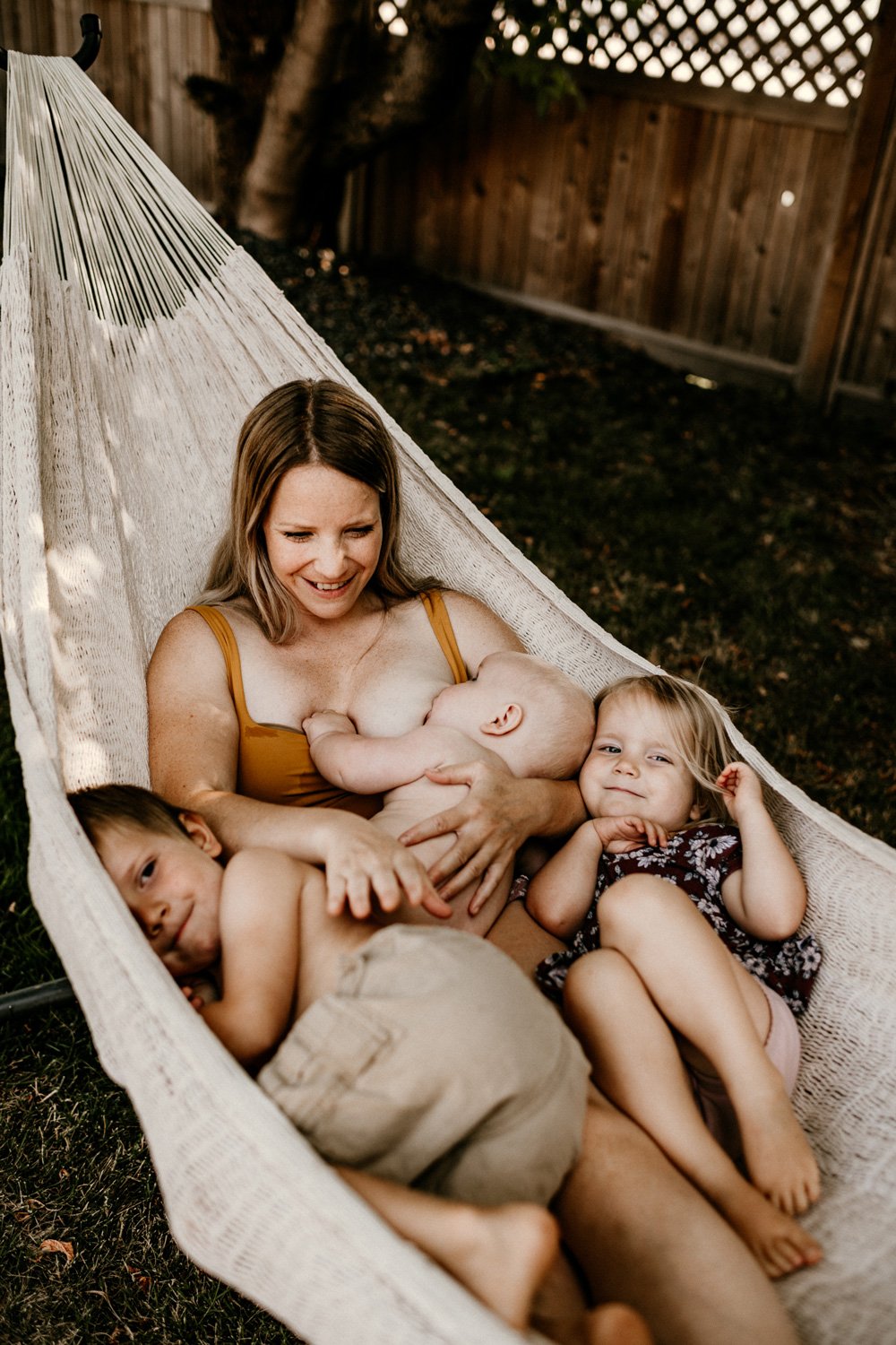A woman with three young children relaxing in a hammock in a backyard. The woman is nursing a baby while two older children cuddle beside her. The setting includes a wooden fence and a tree.