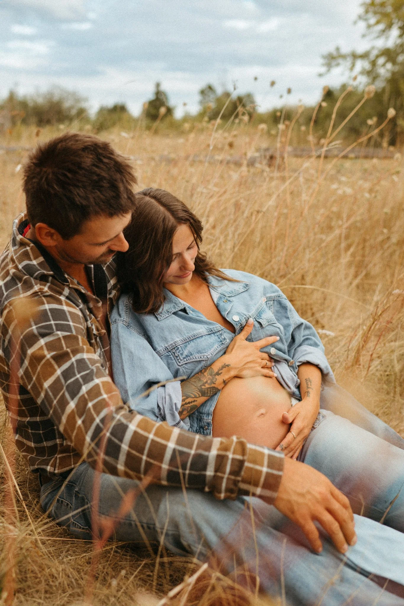A pregnant woman and a man sitting in a field of tall, dried grass, affectionately touching her belly.