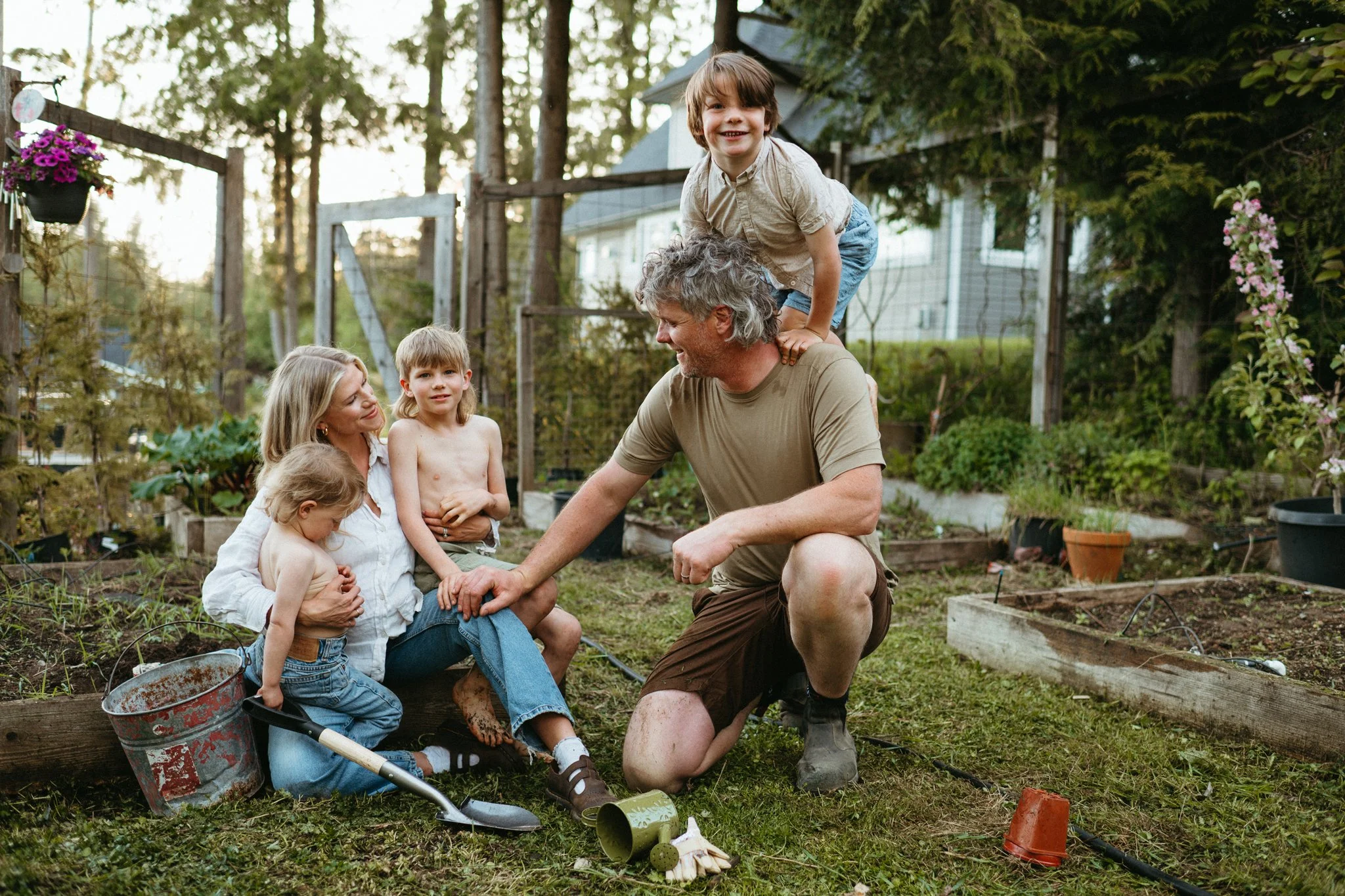 A family gardener working in their backyard garden in the evening, with five children sitting or playing around him.