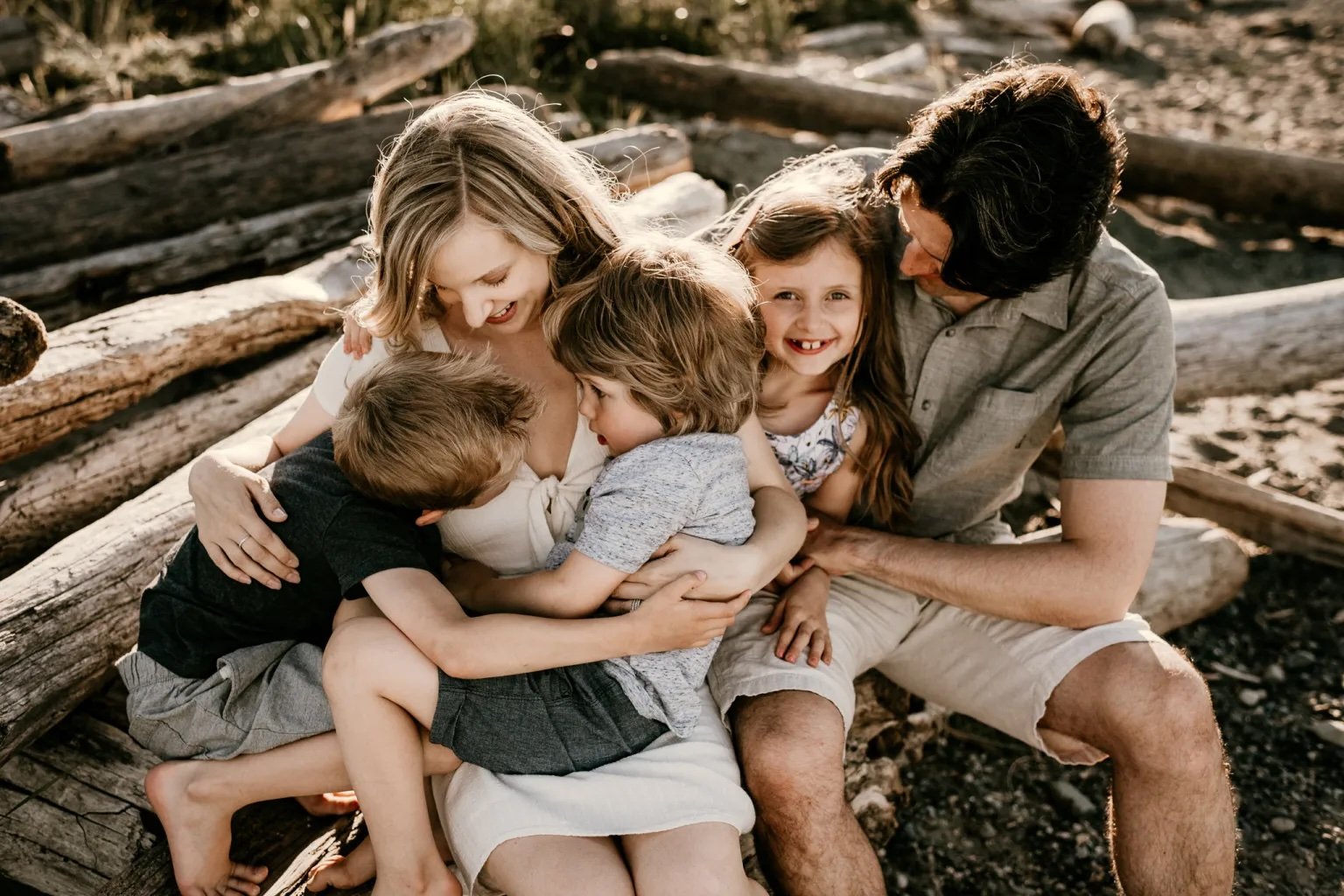 A family sitting together, two adults and three children, embracing and smiling in a natural outdoor setting with wooden logs in the background.