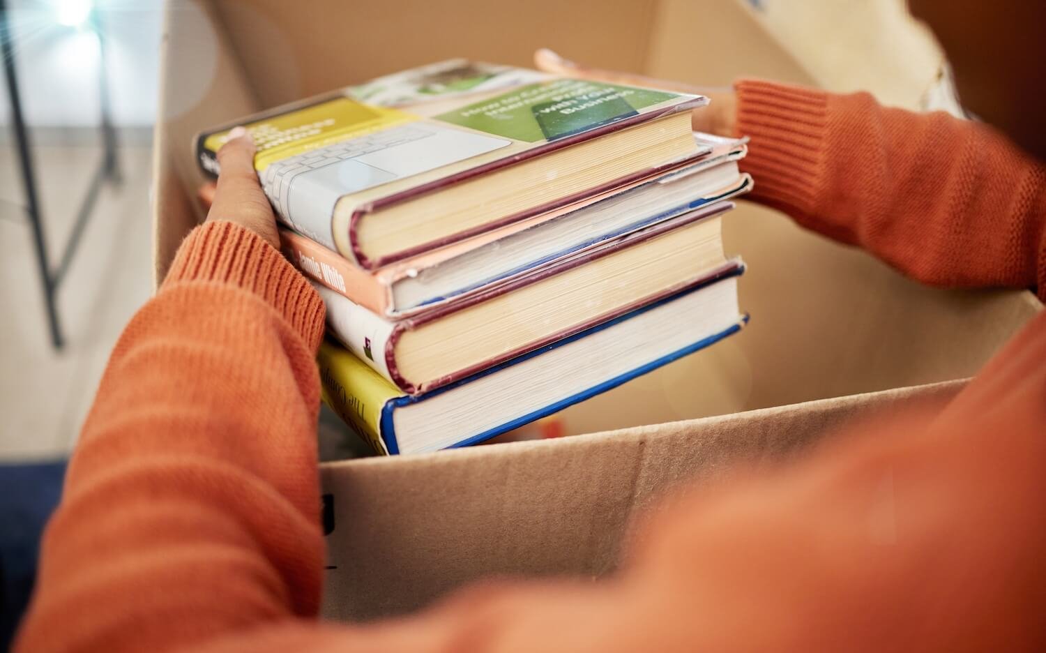 A person wearing an orange sweater places a stack of hardcover books into a cardboard donation box, symbolizing charitable giving and education access.