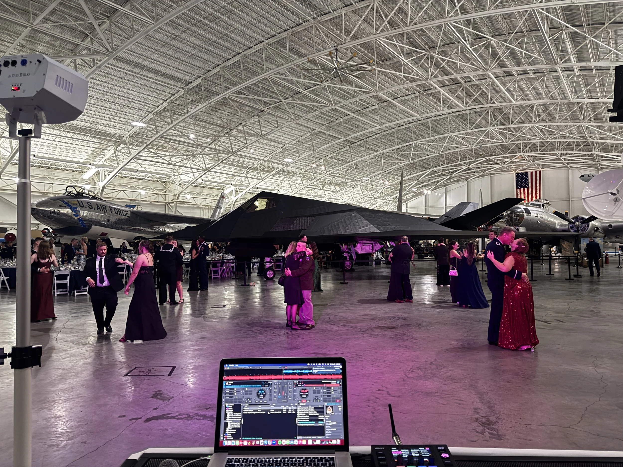 An indoor aviation museum with vintage aircraft, including a U.S. Air Force jet, on display. Guests are attending an event, dressed in formal attire. A DJ setup with a laptop and sound equipment is in the foreground, and an American flag hangs on the