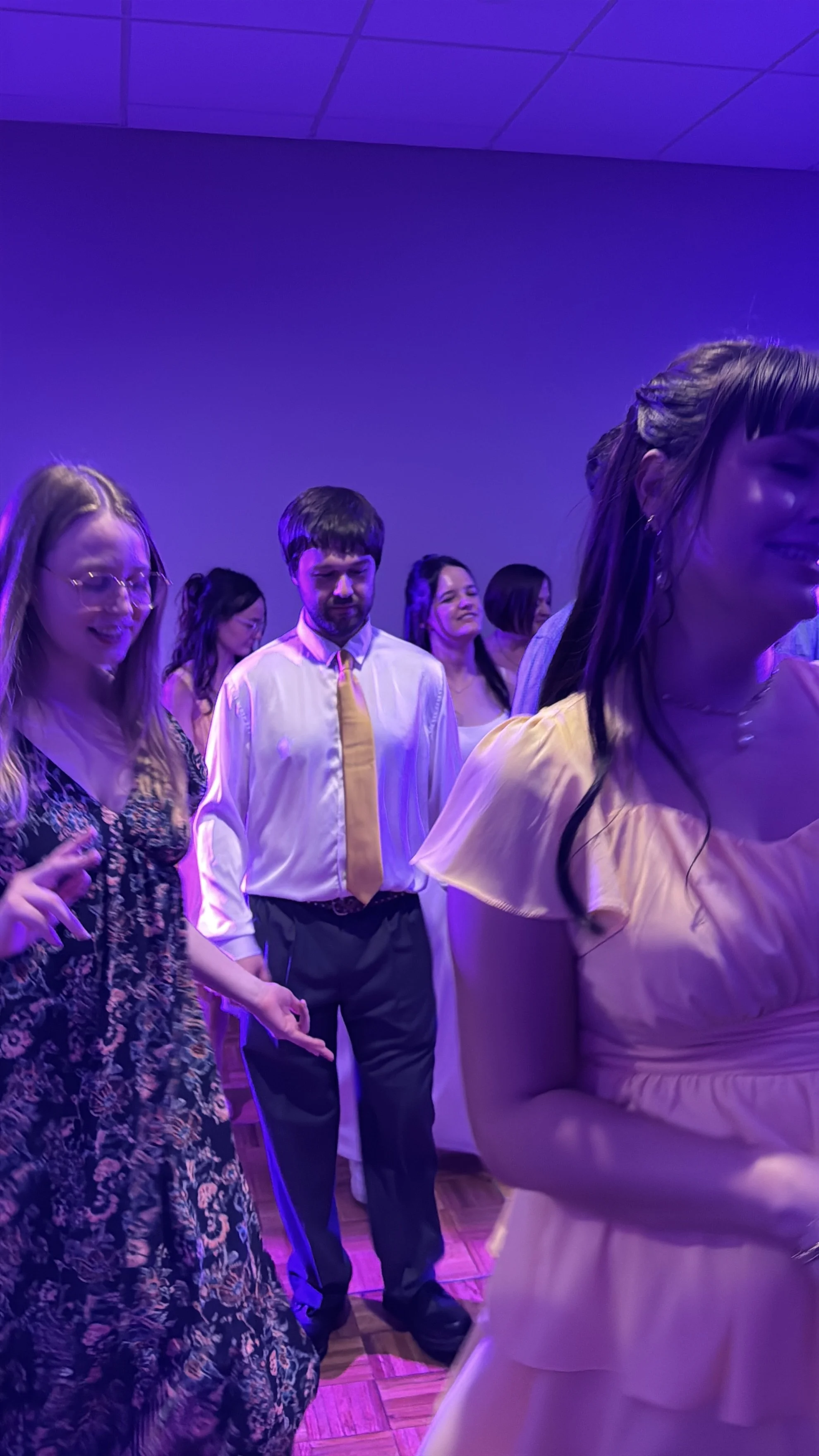A group of young adults dancing together in a room with purple lighting, some with eyes closed and smiling.