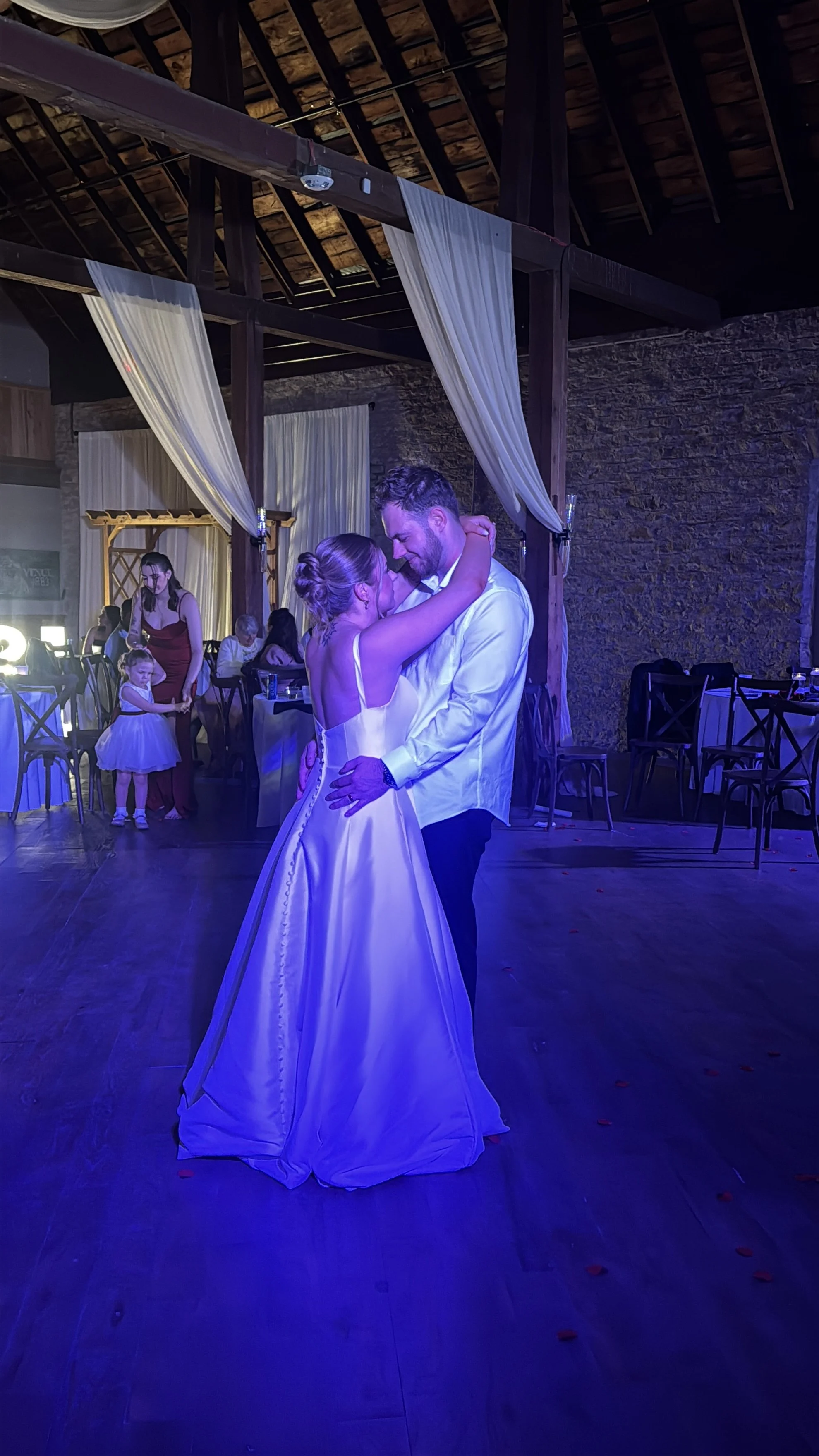 A couple dancing closely at a wedding reception, with guests in the background, some dressed in formal attire, and the bride wearing a white gown.
