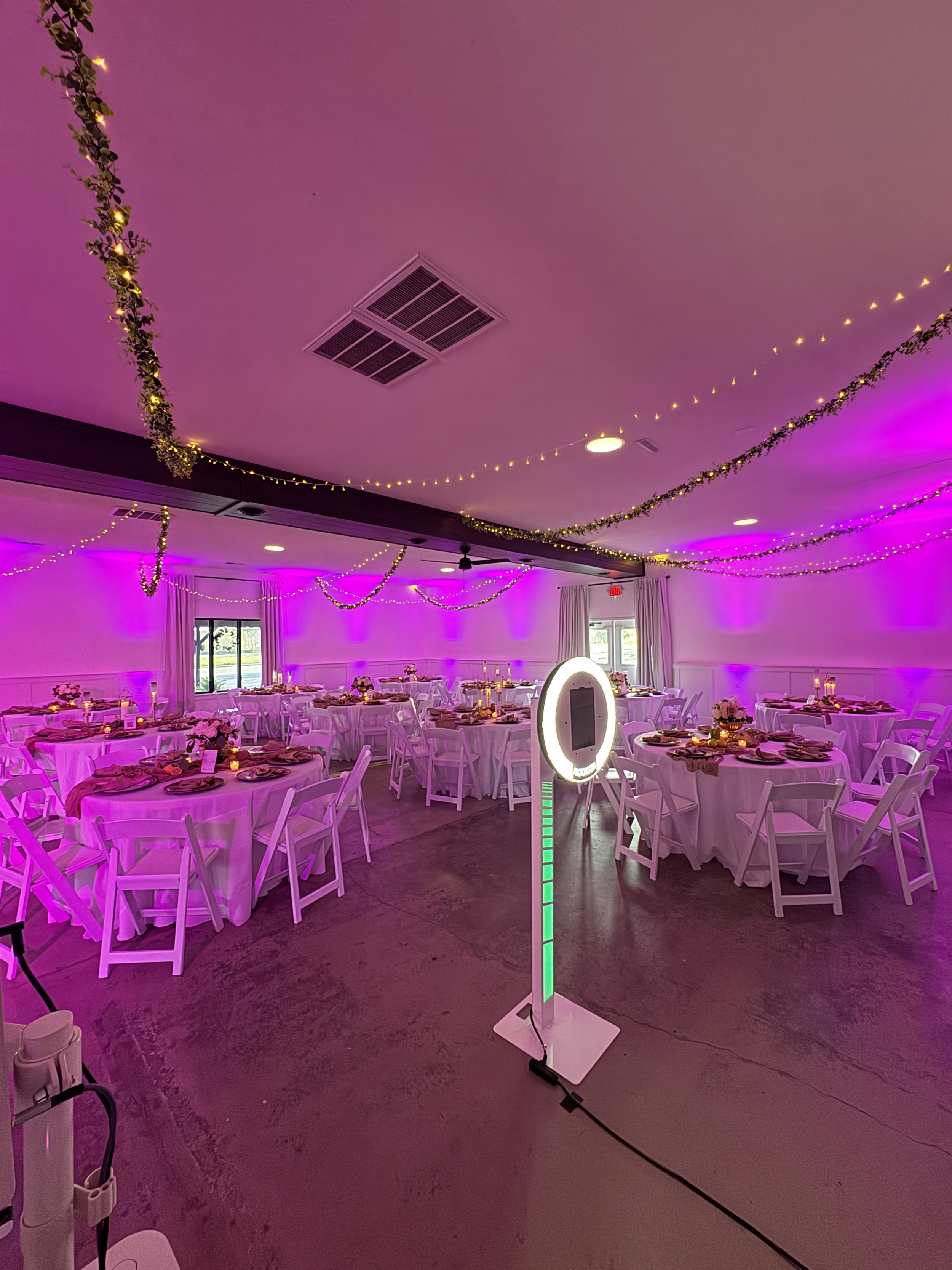 Wedding reception hall decorated with string lights, flowers, and purple uplighting, with round tables set with pink napkins, plates, and floral centerpieces.