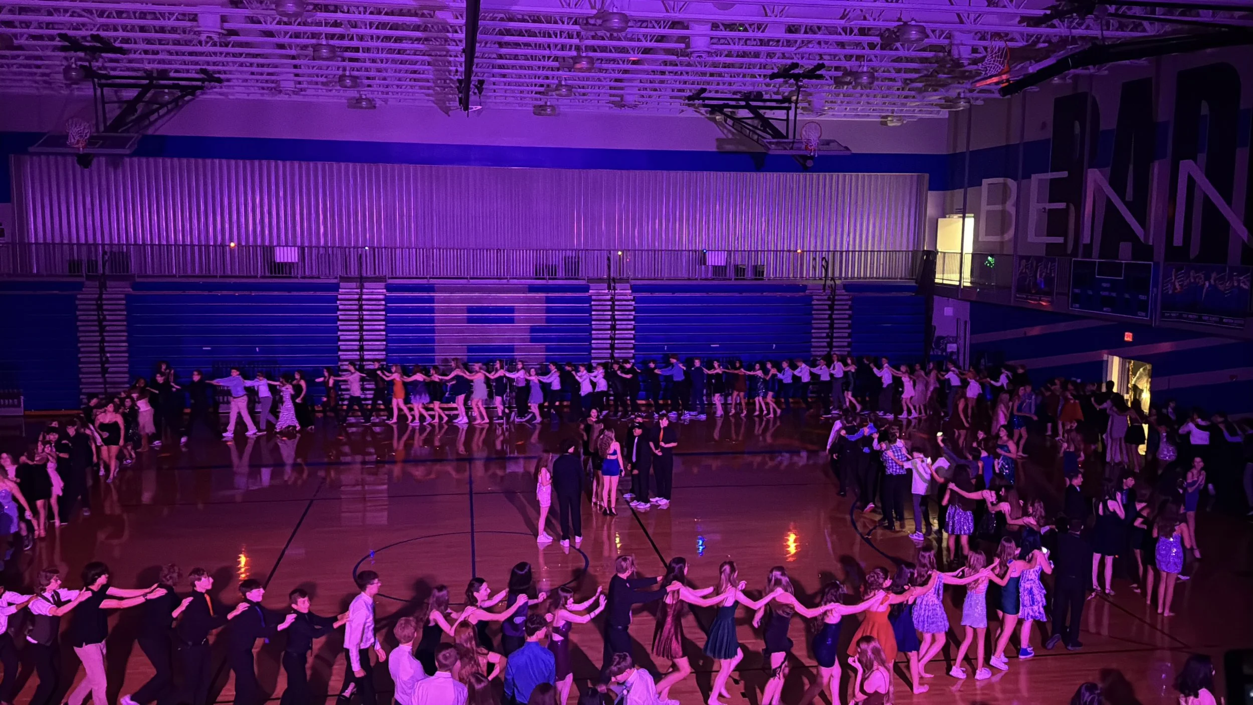Many people are dancing in pairs in a gymnasium, forming a long chain. The gym is illuminated with purple lighting, and the walls display the word 'BENNING.'