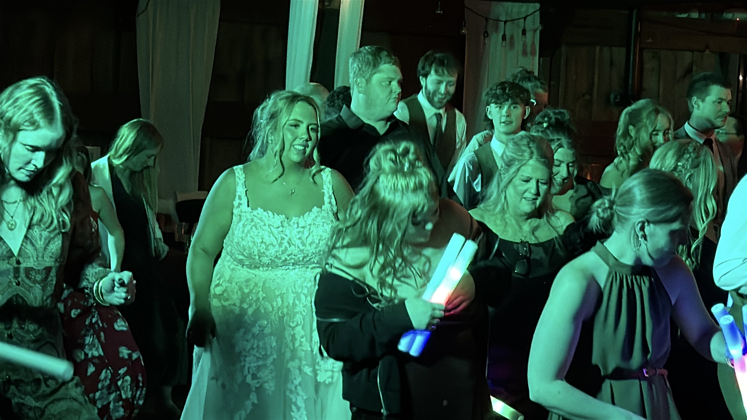 Group of people attending an indoor event, some holding glow sticks, with green and blue lighting, in a rustic wooden venue.