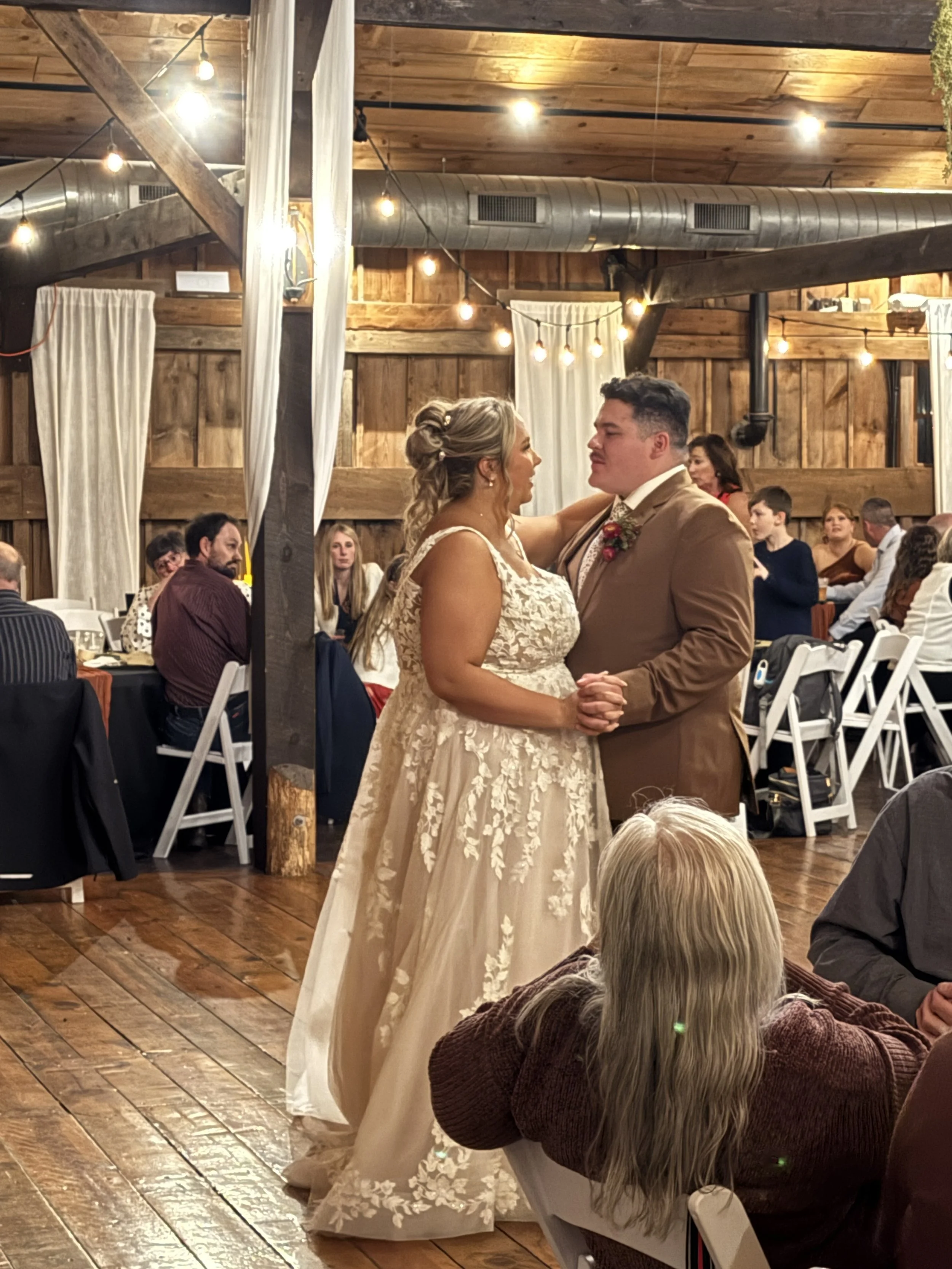 A bride and groom are dancing at their wedding reception inside a rustic barn with wooden walls and ceiling. Guests are seated at tables watching them.