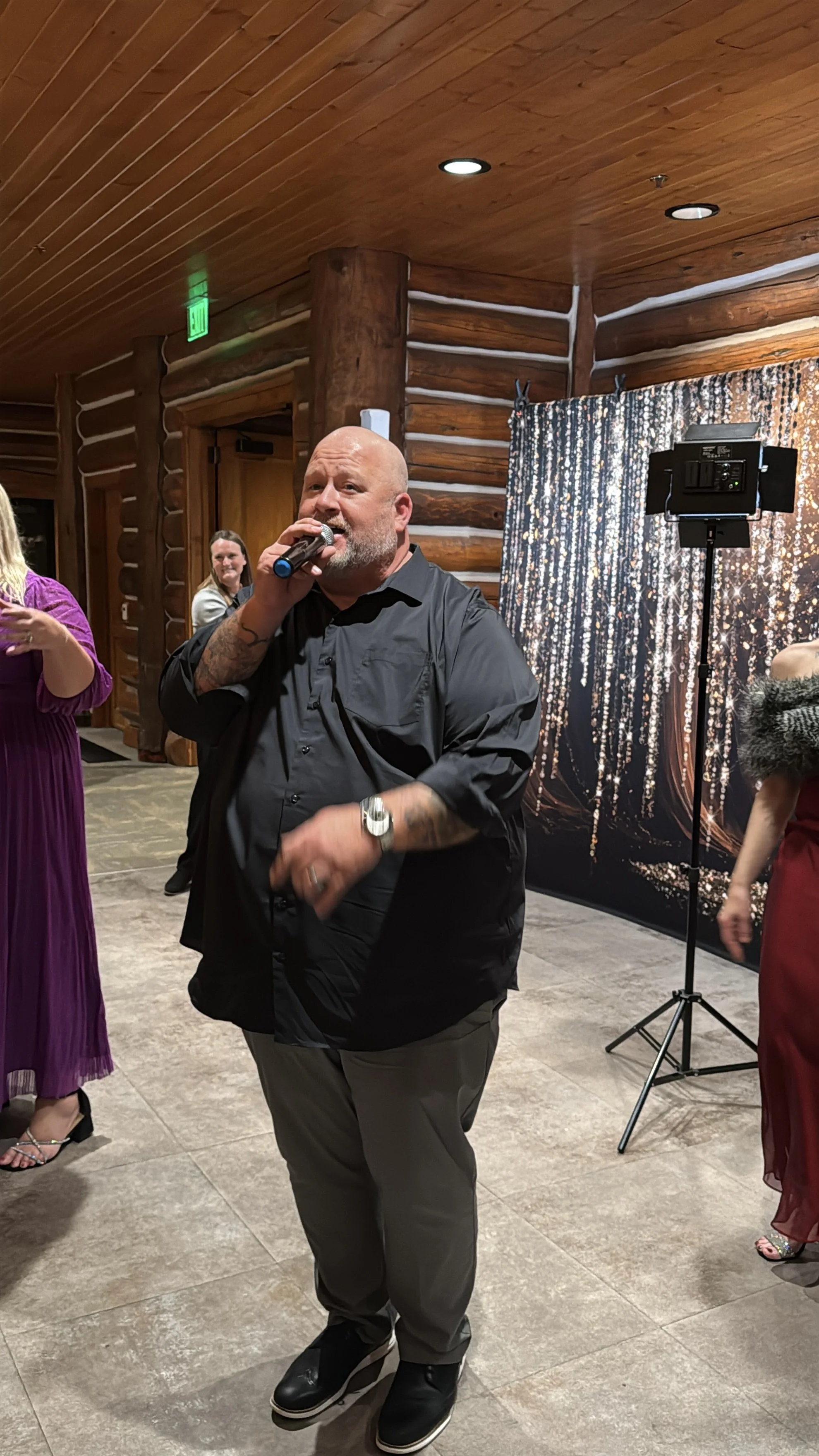 A man with a beard and tattoos singing into a microphone at a party or event, with women in elegant dresses around him, in a wooden log cabin-style room with a glittery backdrop and lighting equipment.