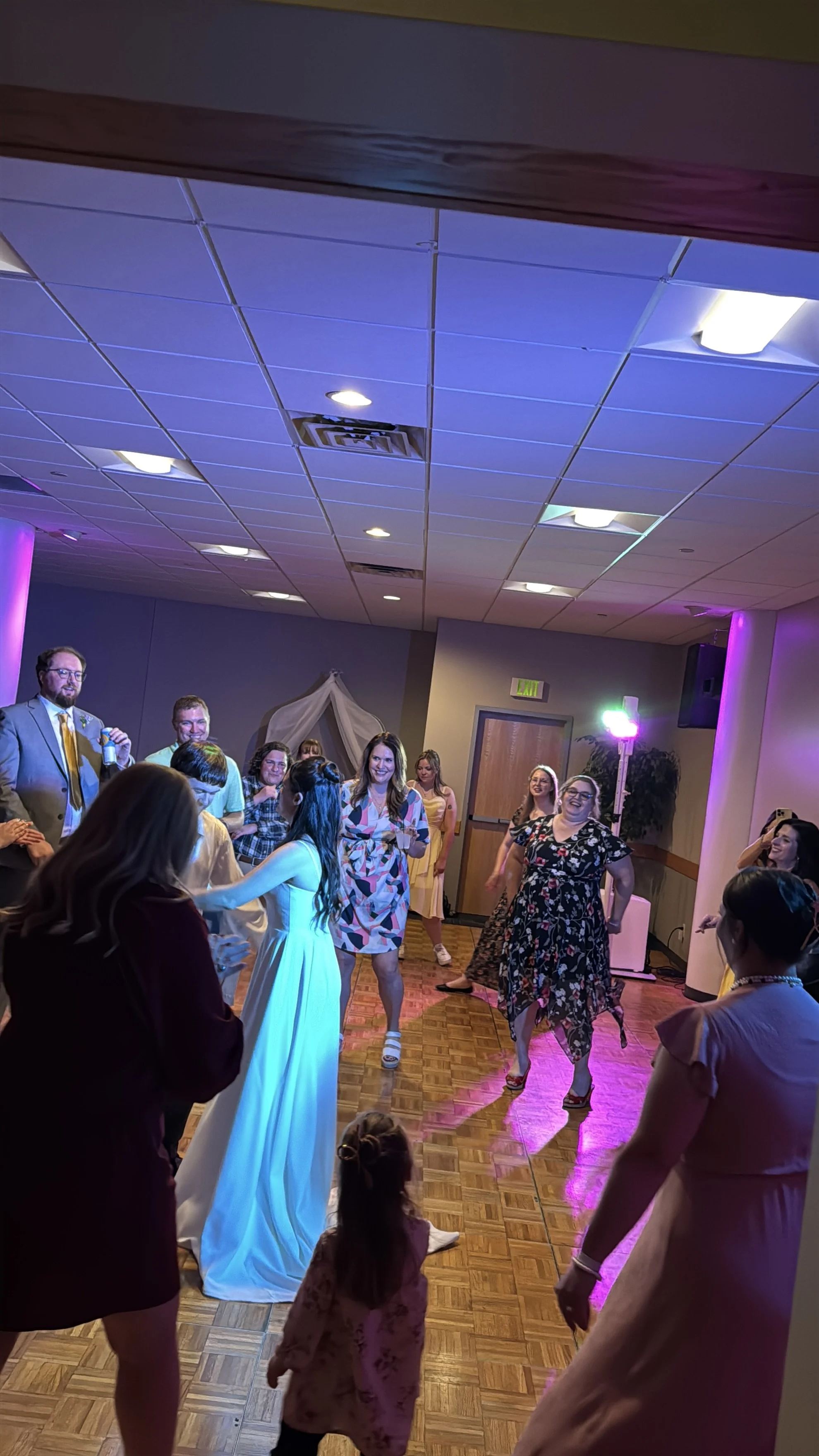 People dancing and socializing at an indoor party or celebration with colorful lighting and a wooden dance floor.