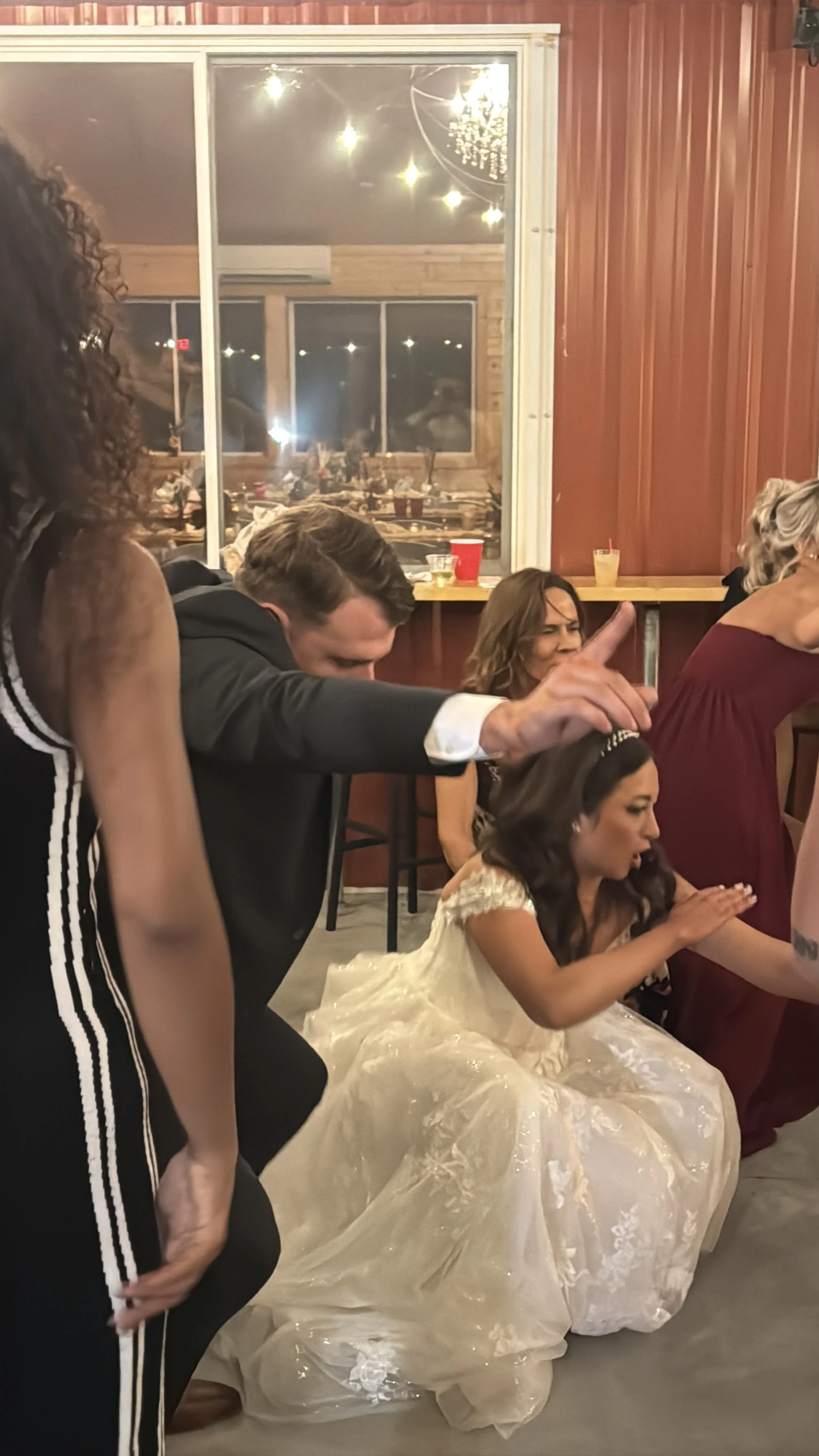 A woman in a white wedding gown sitting on the floor with her hands clapped, surrounded by people at what appears to be a celebration or wedding event. A man in a dark suit is kneeling next to her, and other women are nearby, some with their eyes clo