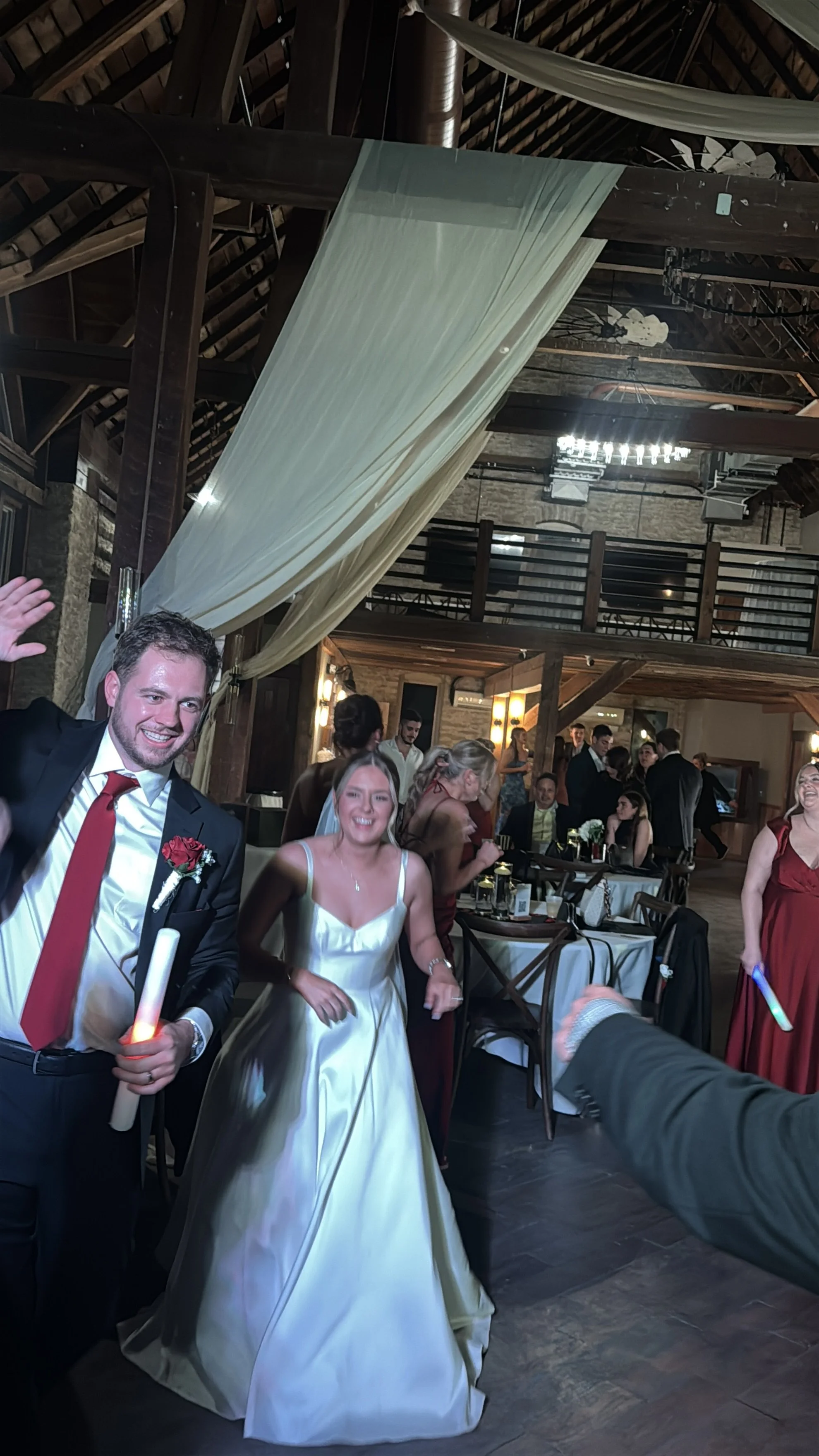People celebrating at a wedding reception in a rustic venue with wooden beams, draped fabric, and a chandelier, with several guests smiling and holding glowing light sticks.
