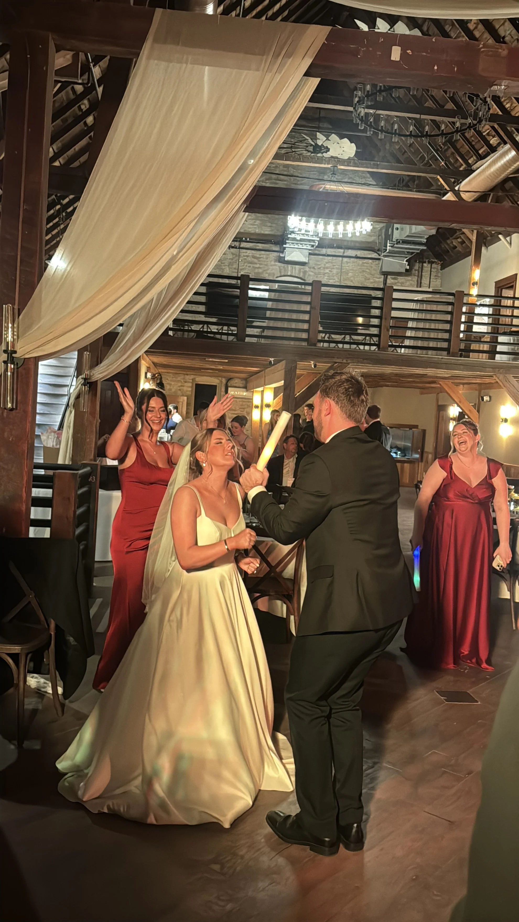 A wedding reception with a bride in a white gown and veil, holding a candle, standing with a groom in a black suit. Two women in red dresses are dancing and smiling in the background. The setting is a rustic hall with wooden beams, chandeliers, and a