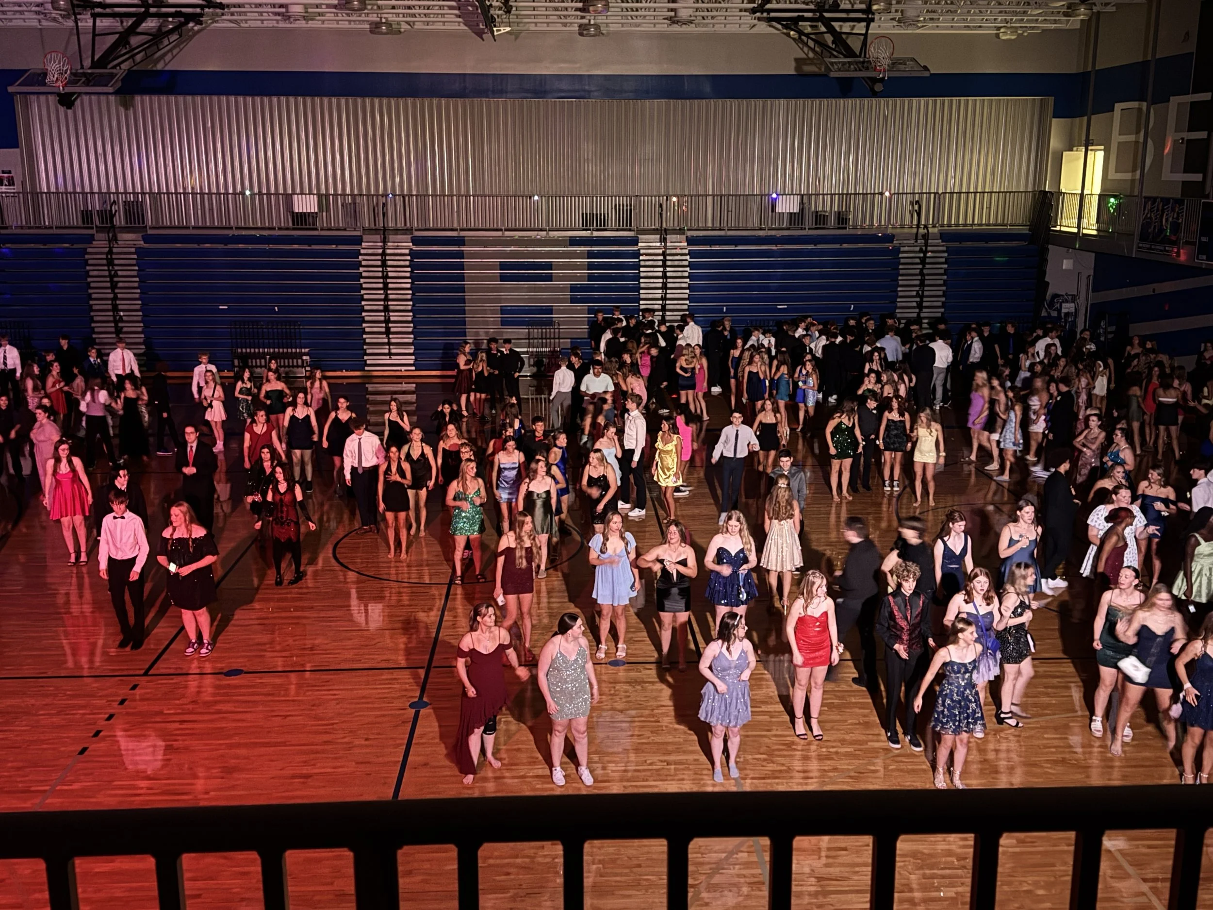 Young people dressed in formal attire gathering in a gymnasium, with some sitting and others standing, as part of a dance or social event.