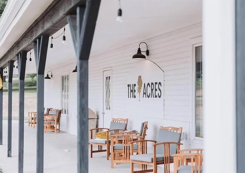 Porch with wooden chairs and string lights, white exterior wall with sign that reads 'The Acres' near a door and window.