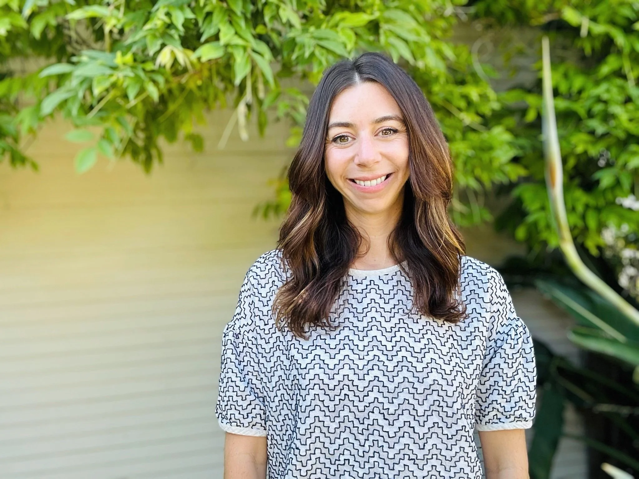 Smiling woman standing outside with greenery background, wearing a patterned shirt.