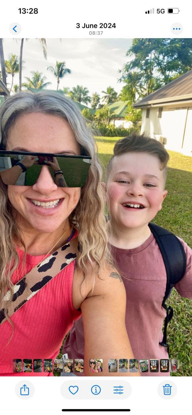 Woman with long gray hair wearing large sunglasses and a red top, smiling, taking a selfie with a smiling boy with short hair, wearing a pinkish shirt and backpack, outdoors in a tropical area with palm trees and houses.