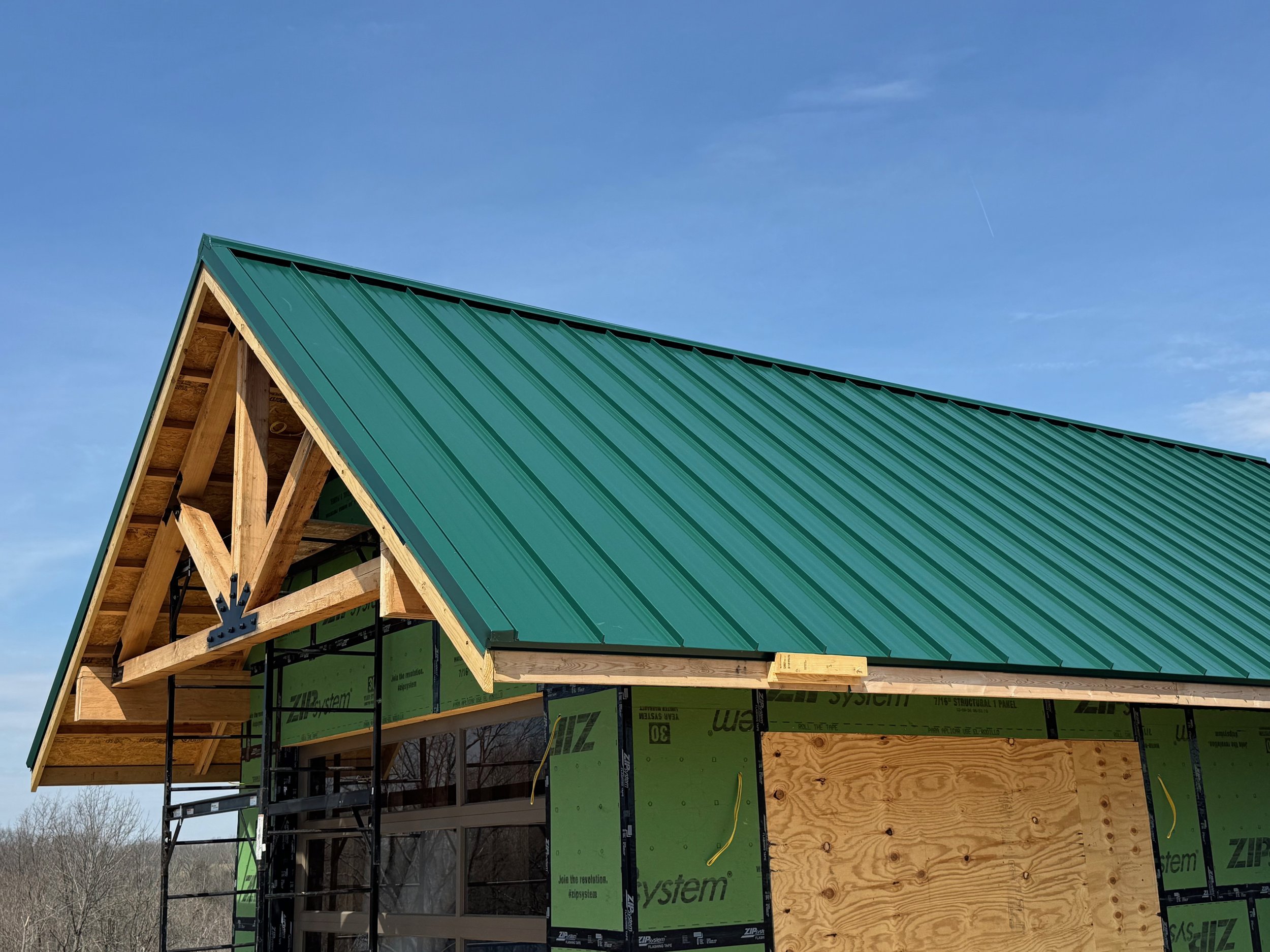 Partially constructed house with green metal roof, plywood siding, and scaffolding, under a blue sky.