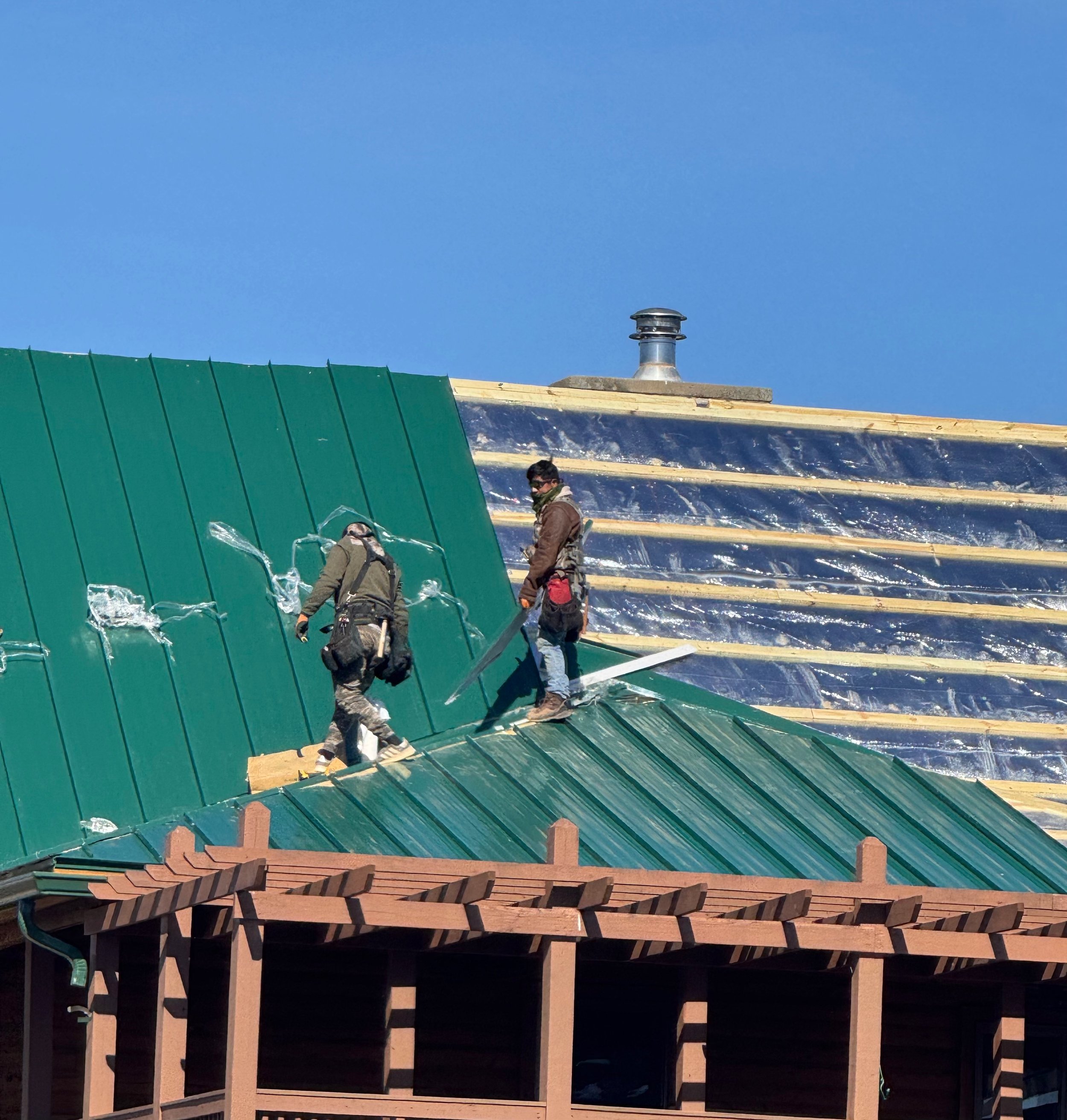 Two construction workers installing green metal roofing on a house under a clear blue sky.