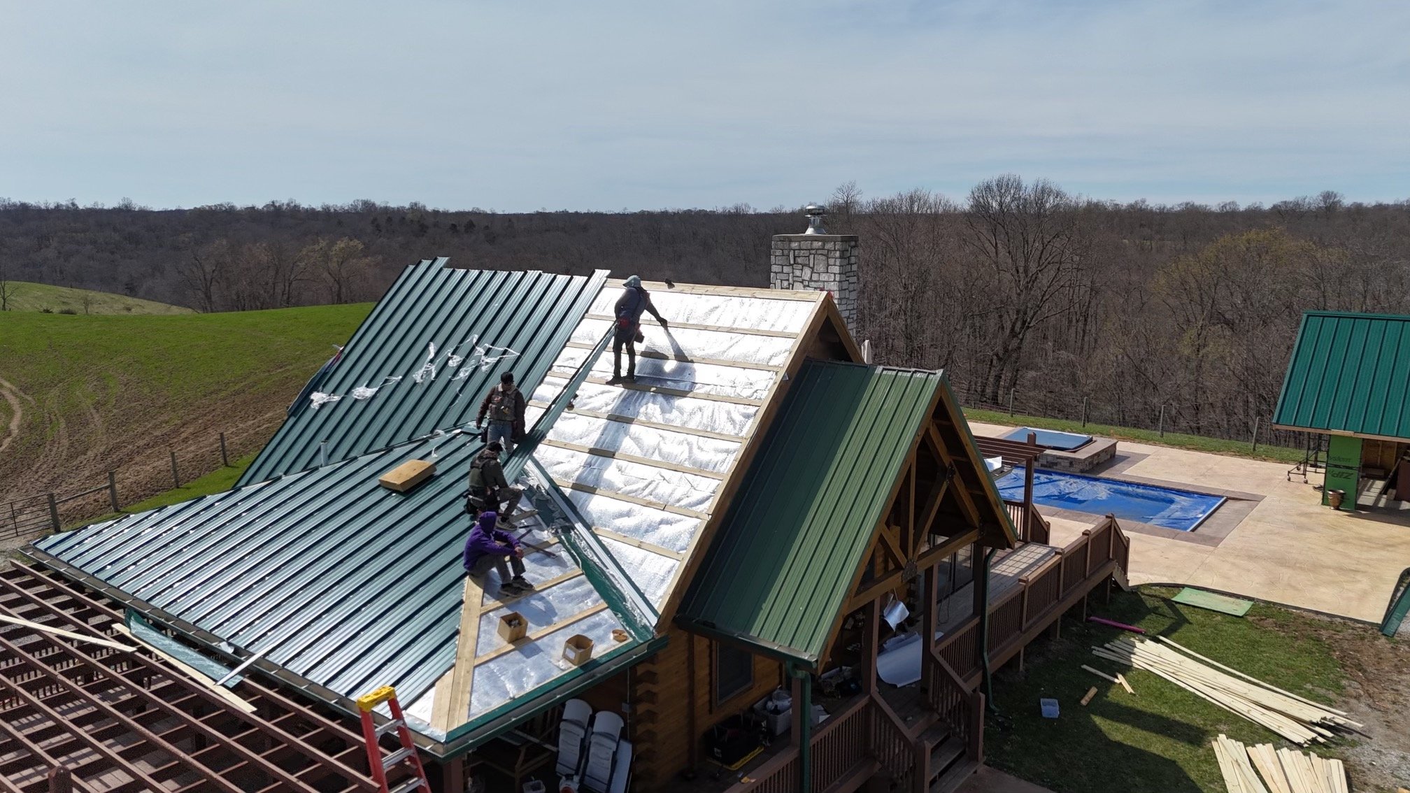 Construction workers installing a metal roof on a house with a wooden deck, in a rural area with trees and a swimming pool in the background.