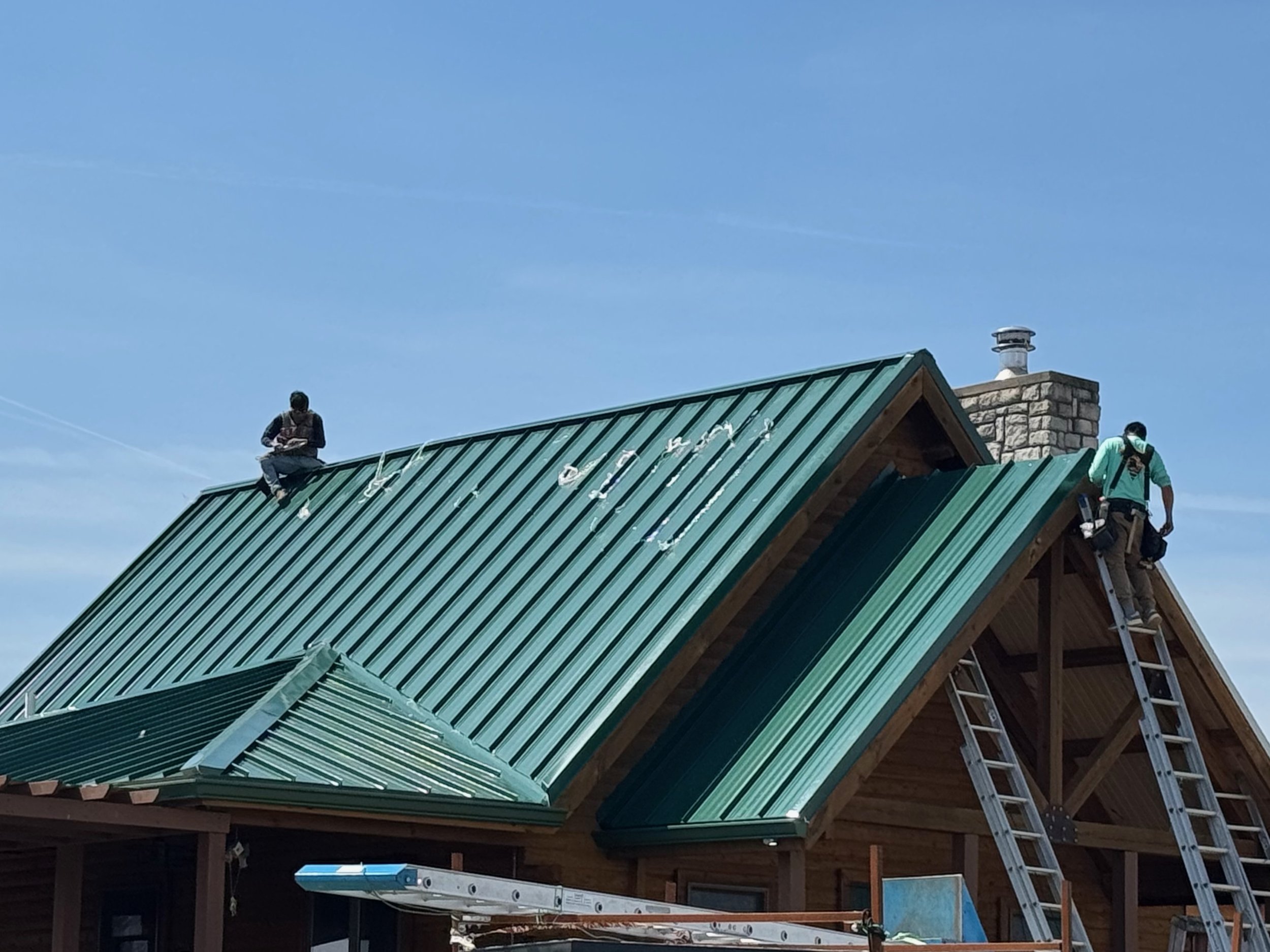 Two workers installing or repairing a green metal roof on a wooden house, with ladders leaning against the roof and a chimney in the background.