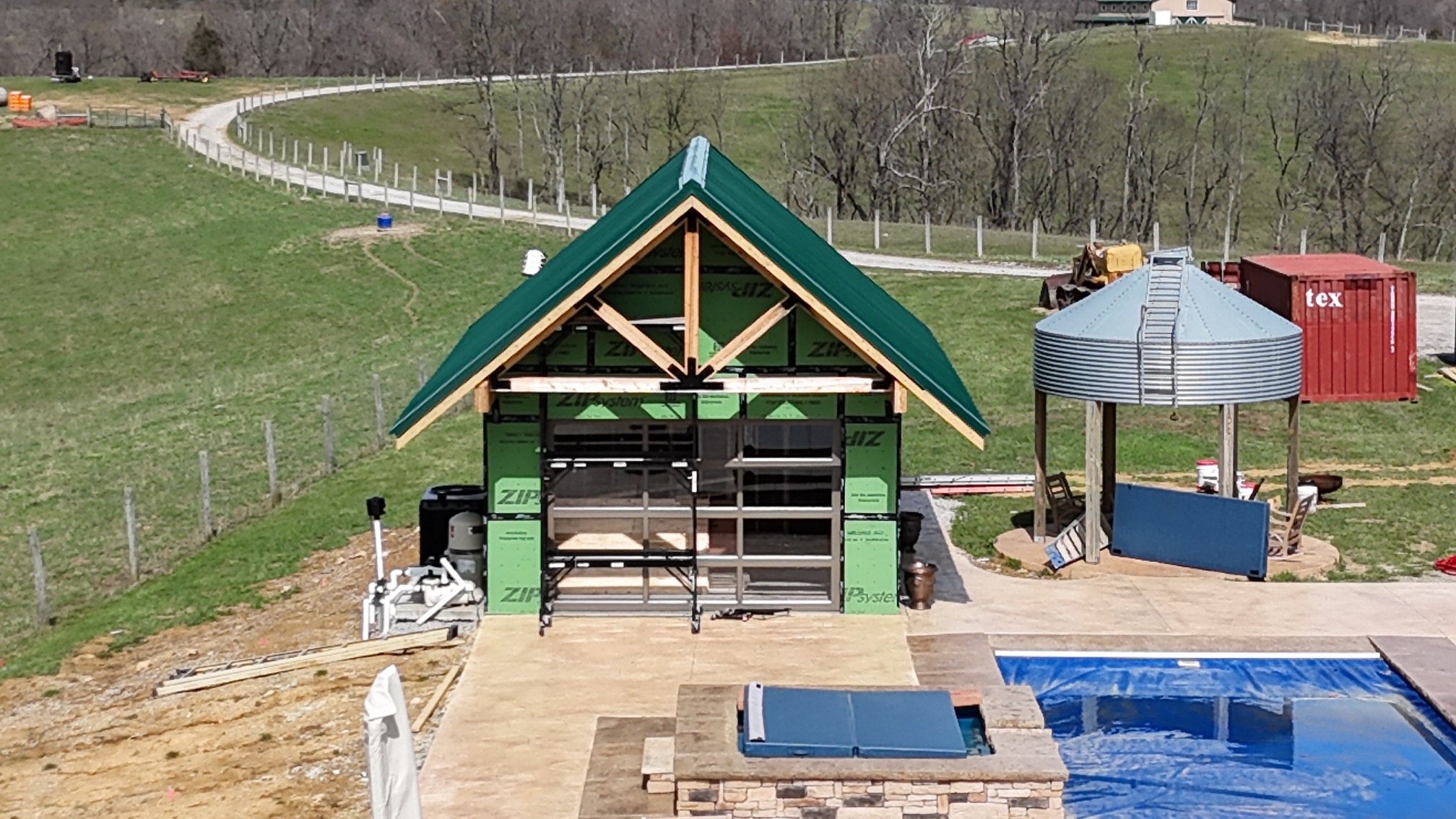 A house under construction with a green metal roof and unfinished exterior, next to a round metal silo, with a pool and construction materials nearby, in a rural setting with grassy hills and trees.