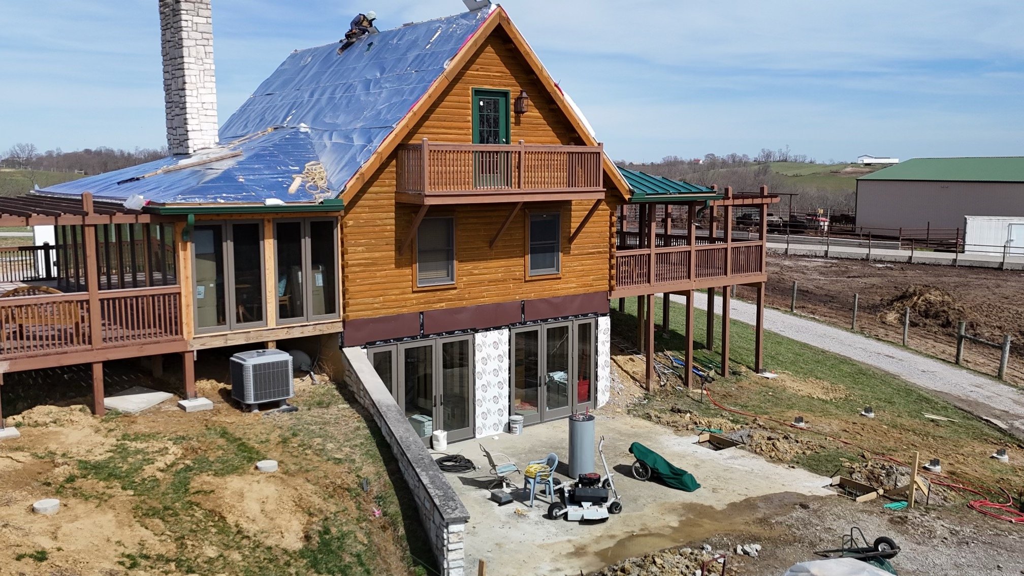 A house under construction with a wood exterior, multiple decks, and a pitched roof in progress, located in a rural area with open land and some construction materials and equipment around.