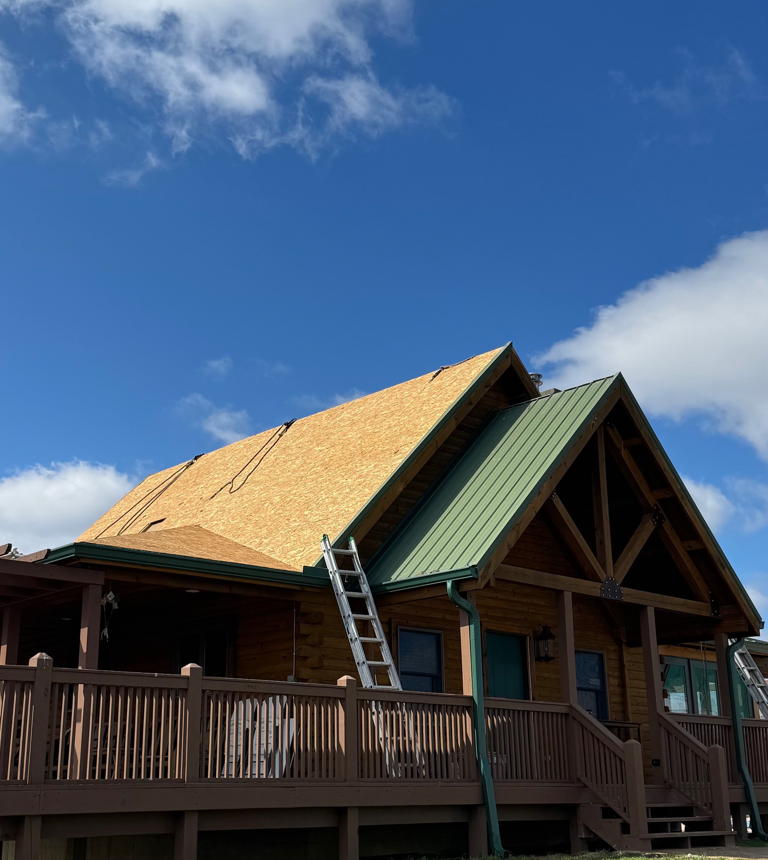 A house under construction with part of the roof covered in new plywood and another part with green metal roofing, a ladder leaning against the house, and a clear blue sky with a few clouds in the background.