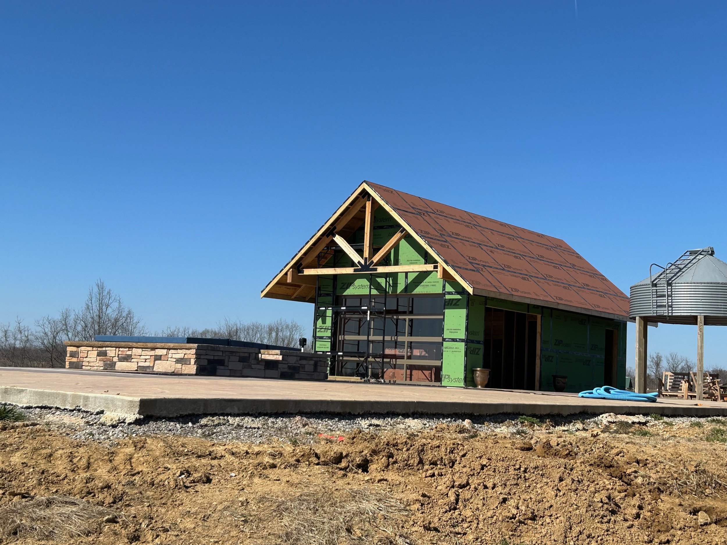 A house under construction with a pitched roof, green sheathing, scaffolding, and a large window opening, on a concrete foundation, with a clear blue sky in the background.