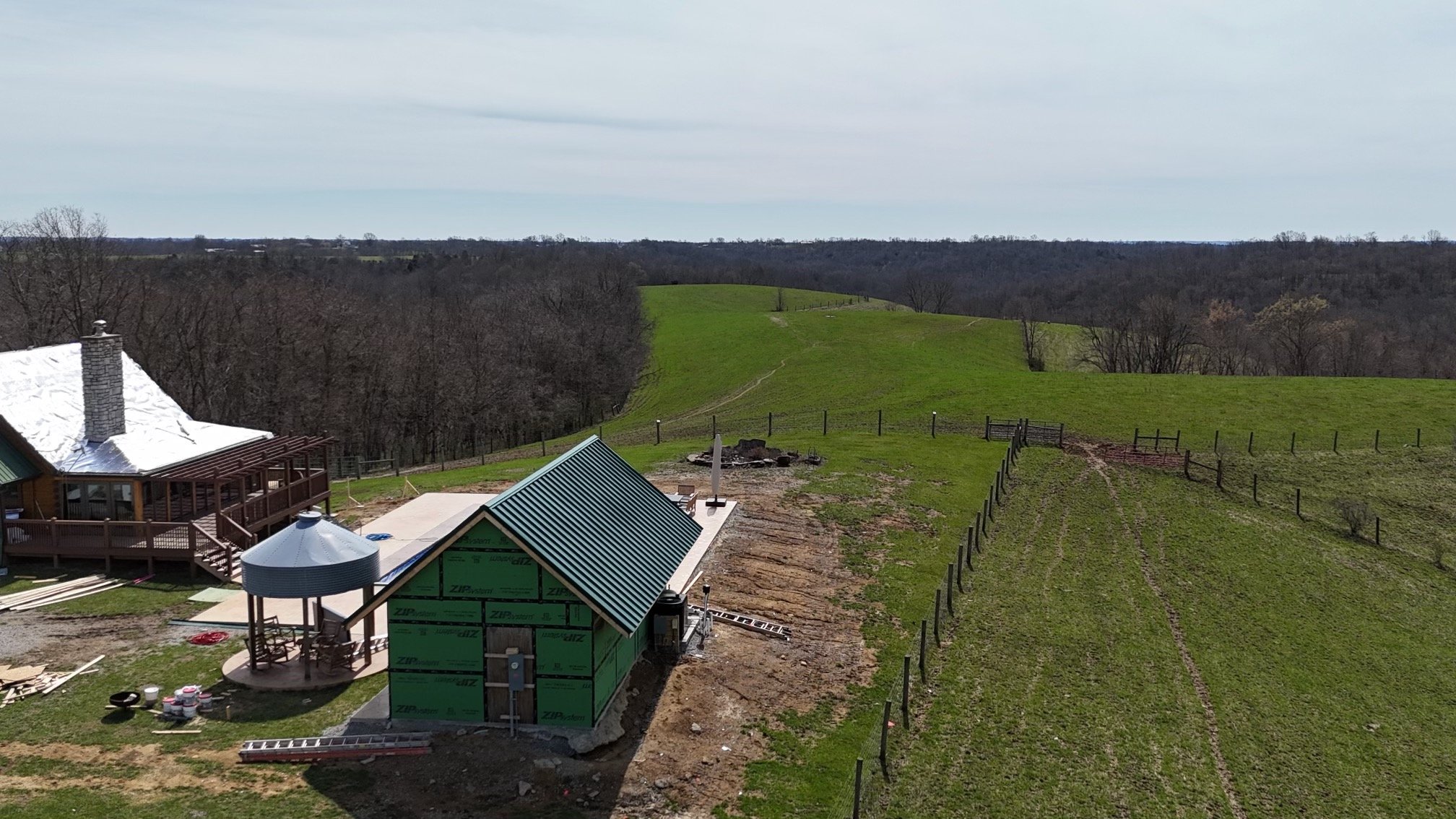 A house under construction on a hillside with a deck, surrounded by green fields and trees, and a distant horizon with a cloudy sky.