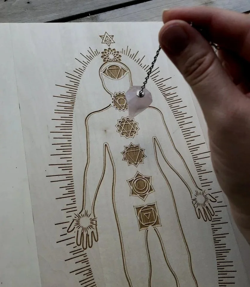 A person holding a pendulum over a wooden chart of the human body with chakra symbols aligned along the body's center, surrounded by a detailed scale.