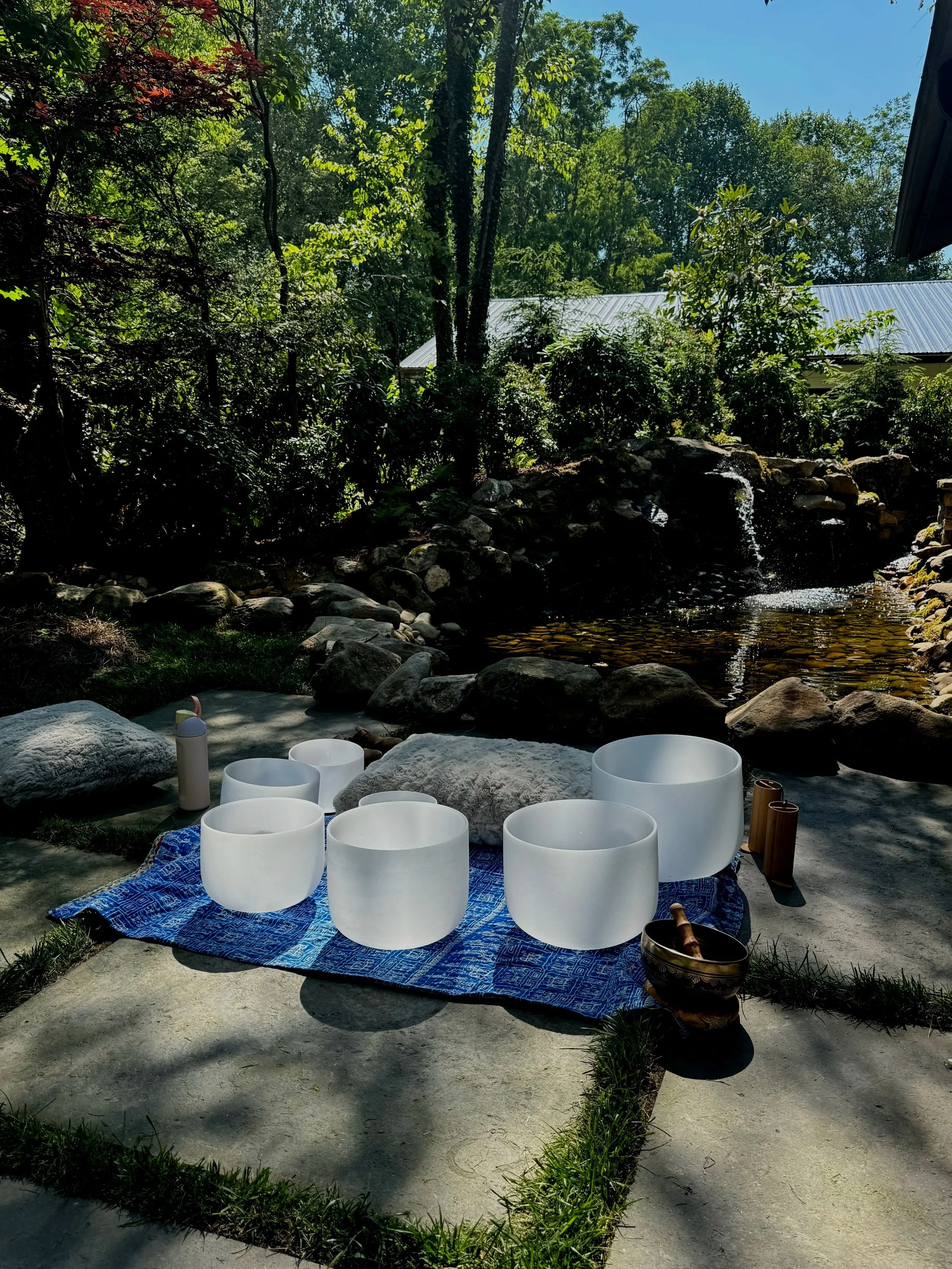 Outdoor setting with six white singing bowls on a blue mat, surrounded by rocks, trees, and a small pond with a waterfall, under a bright sky.