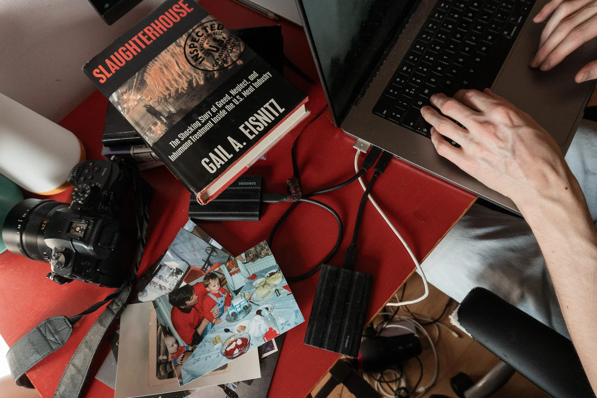 Documentary props and tools, laying on Jack’s red desk. A camera, a stack of old photos, a book with a title ‘Slaughterhouse’ , memory drives and a computer.