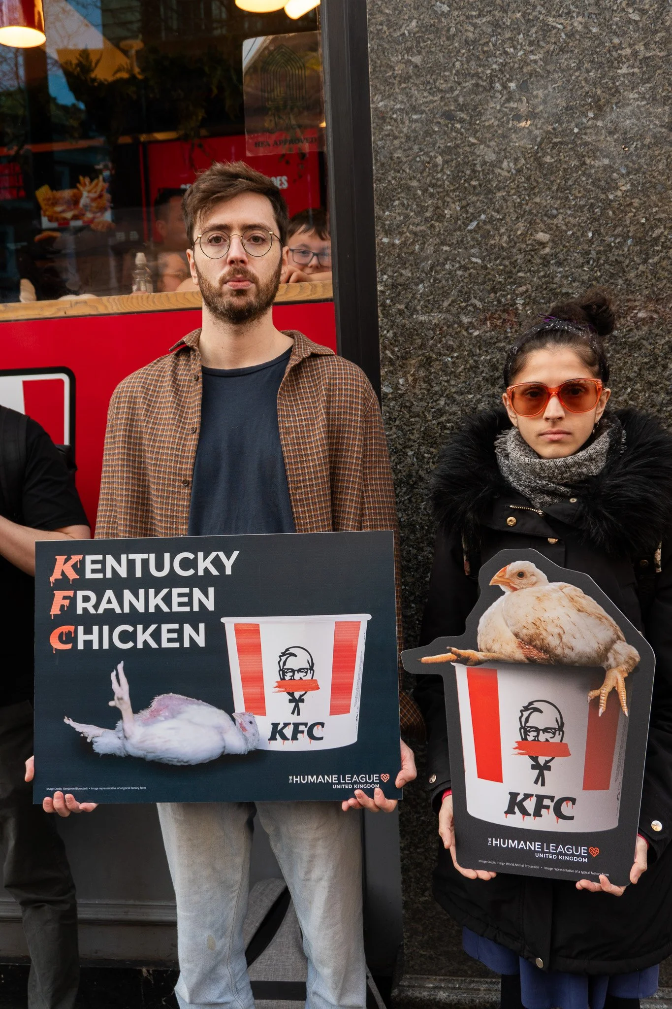 Jack is holding a sign that says  'Kentucky Franken Chicken' and shows an image of a collapsed chicken. Next to him is a woman holding a similar sign. They are protesting in front KFC restaurant.