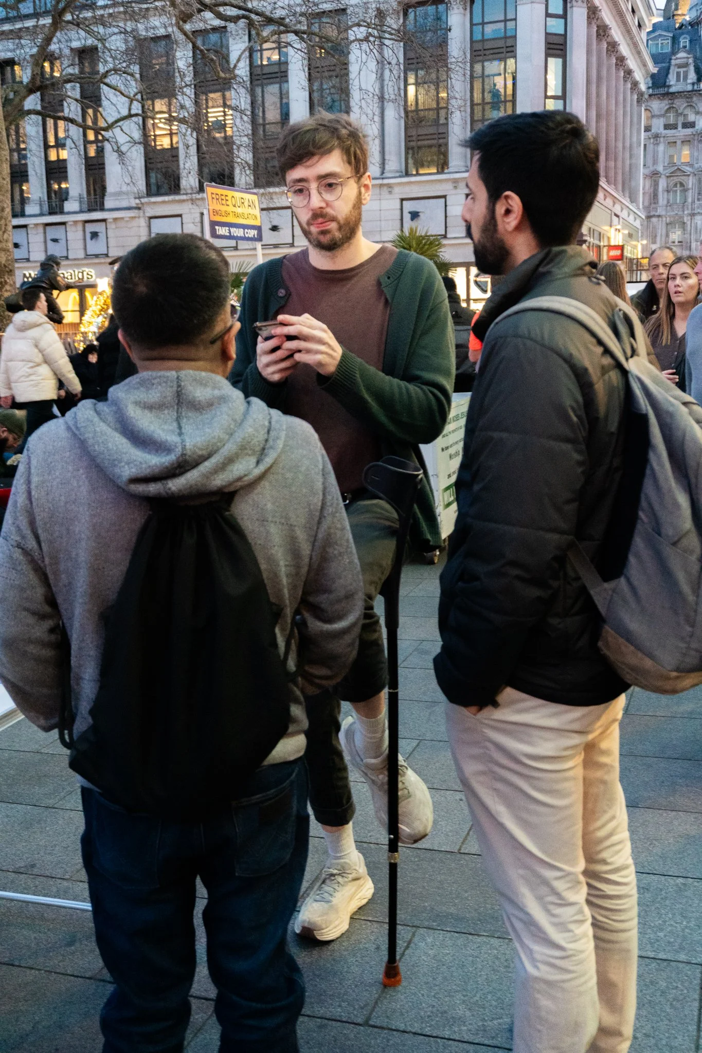 Jack is holding his leg on his crutch and talking to two passersby and asking them to sign a petition on his phone.