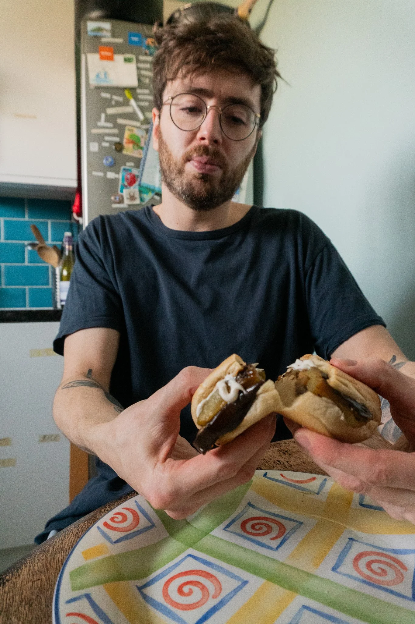 Jack is sitting in a kitchen and breaking a plant-based sandwich into two halves.