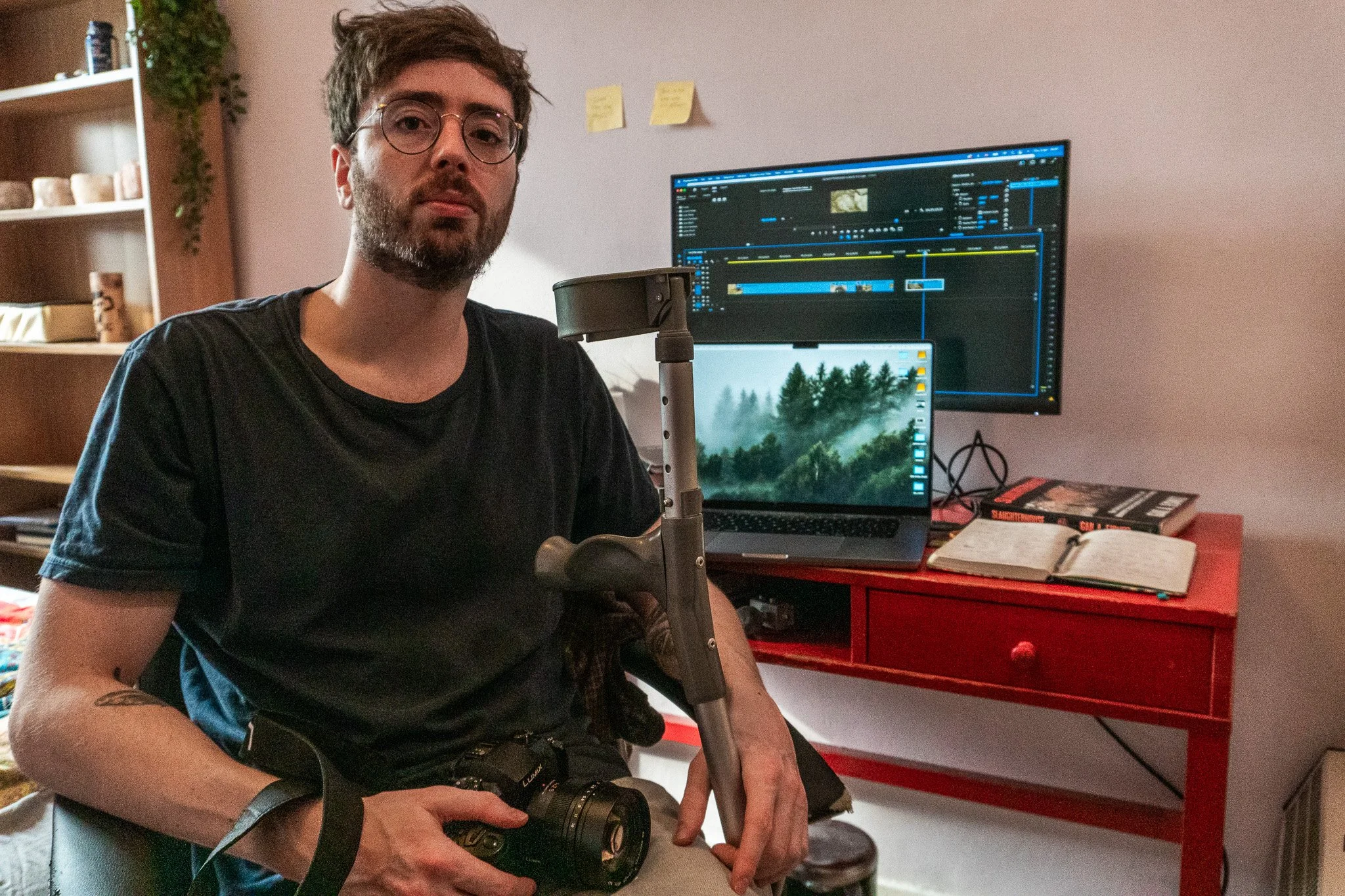 Introduction to Jack Hancock-Fairs.  A 30 year old man with brown hair, beard and round glasses. Jack is sitting in front of his desk, facing the camera holding his crutch and camera.