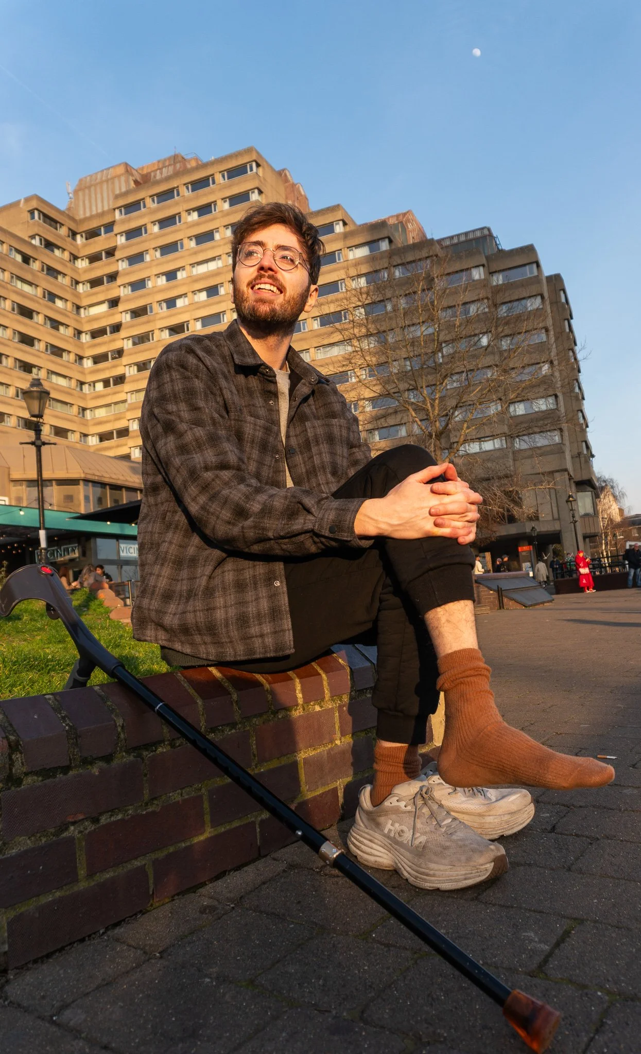 Jack is sitting down to take a break from walking. His crutch is laying beside him. Jack removed one of his legs from a shoe and is hugging it, whilst sitting cross-legged. He is looking up, enjoying the sunny weather.