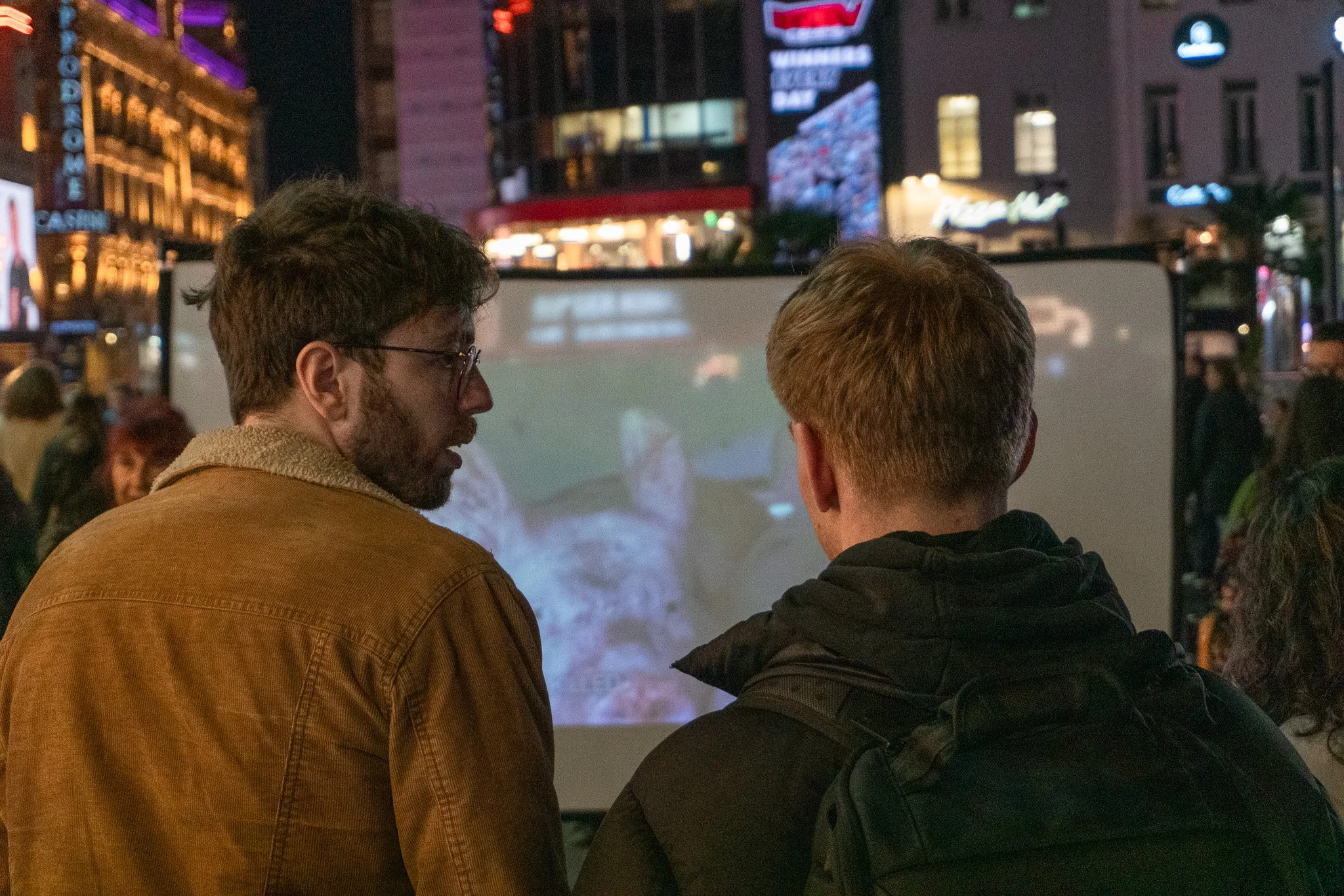 Jack is looking at a man standing beside him, who is looking at a big screen that is showing a close-up of a pig. Jack is educating this man about pig slaughter methods used in the UK.