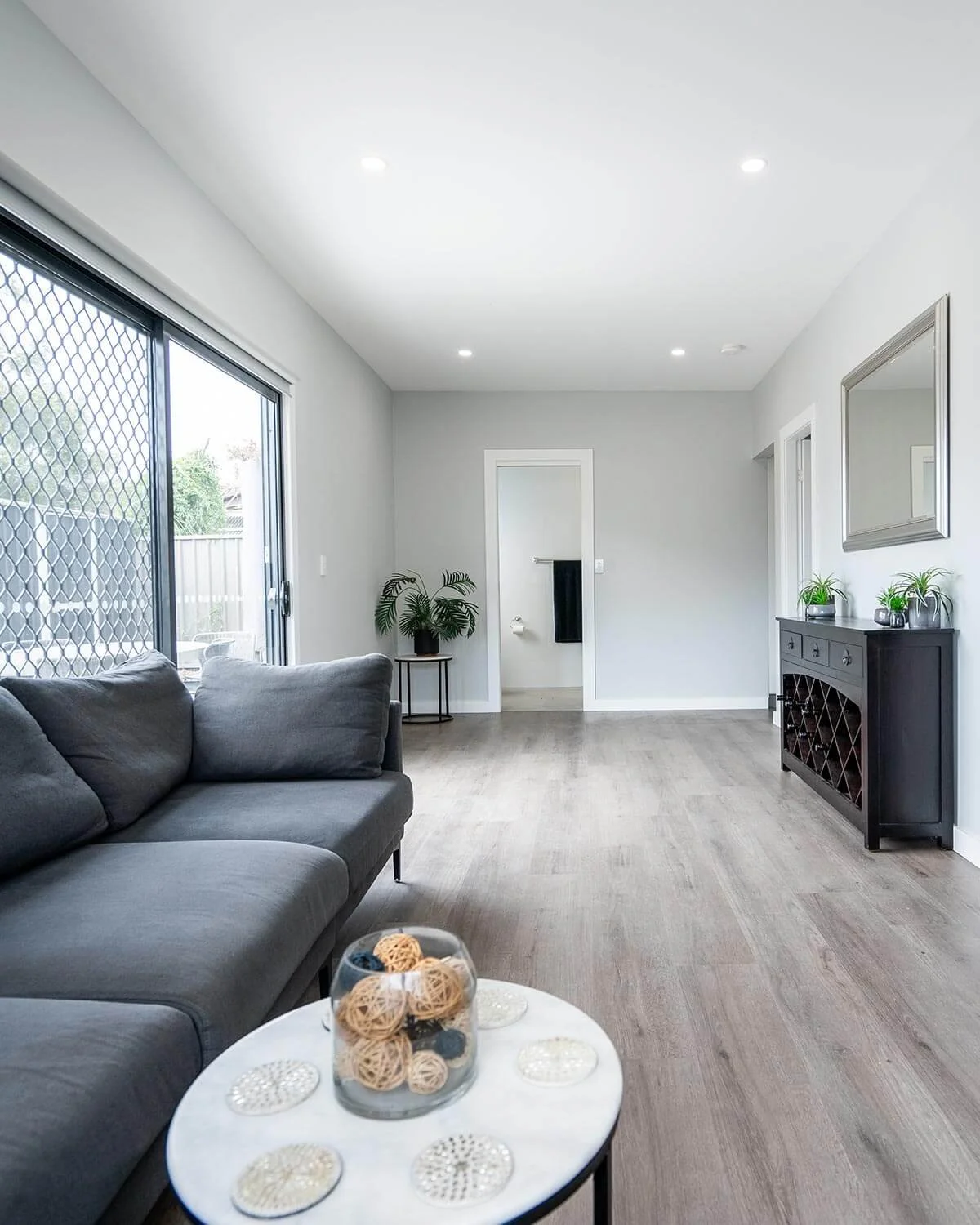 A spacious living room with a gray couch, a marble coffee table with decorative balls, large sliding glass doors with a security screen, a black cabinet with plants, and a mirror on the wall.